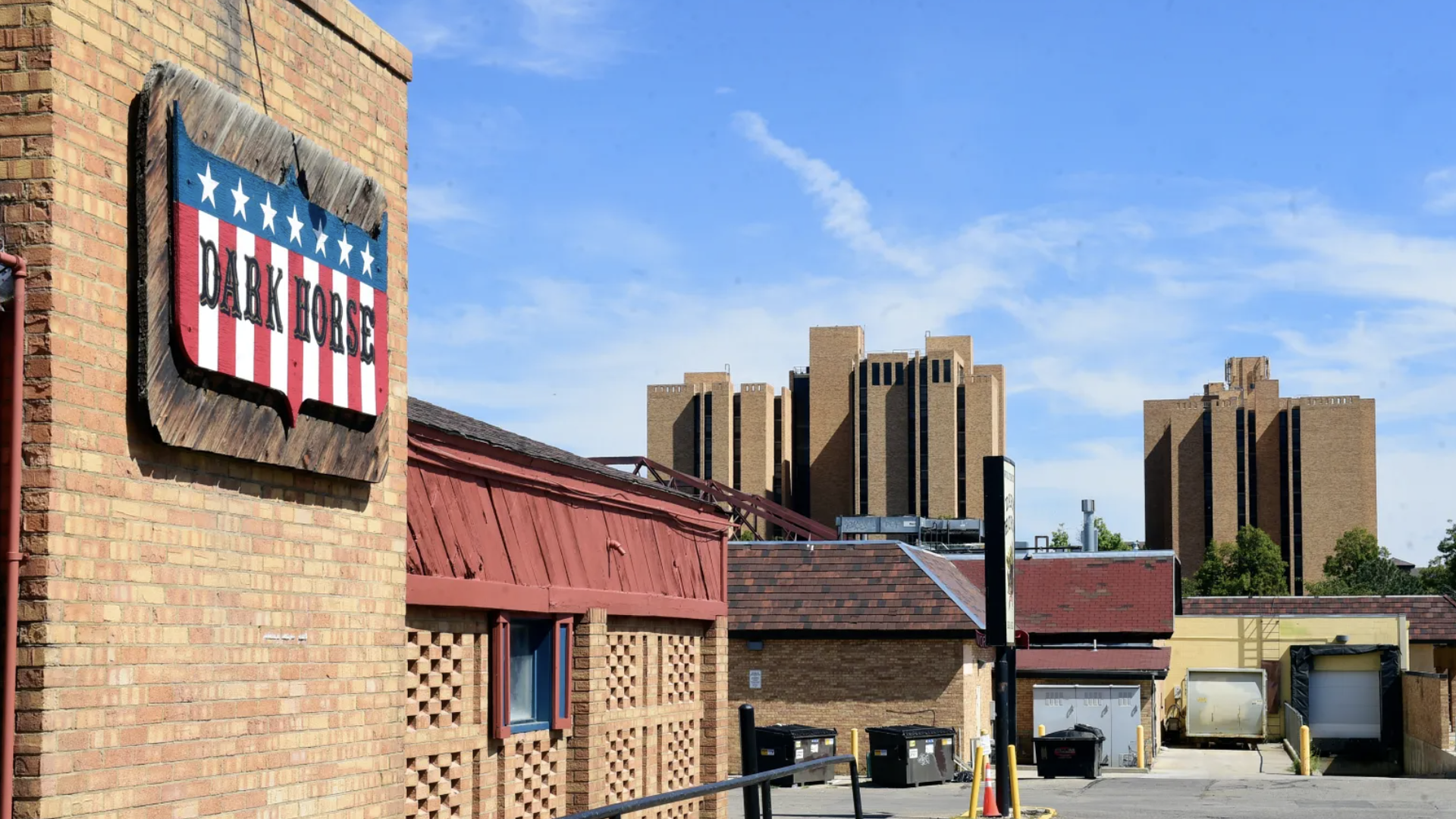 Brick building with a red, white, and blue "Dark Horse" sign, an alley with dumpsters, and tall brown buildings under a clear blue sky.