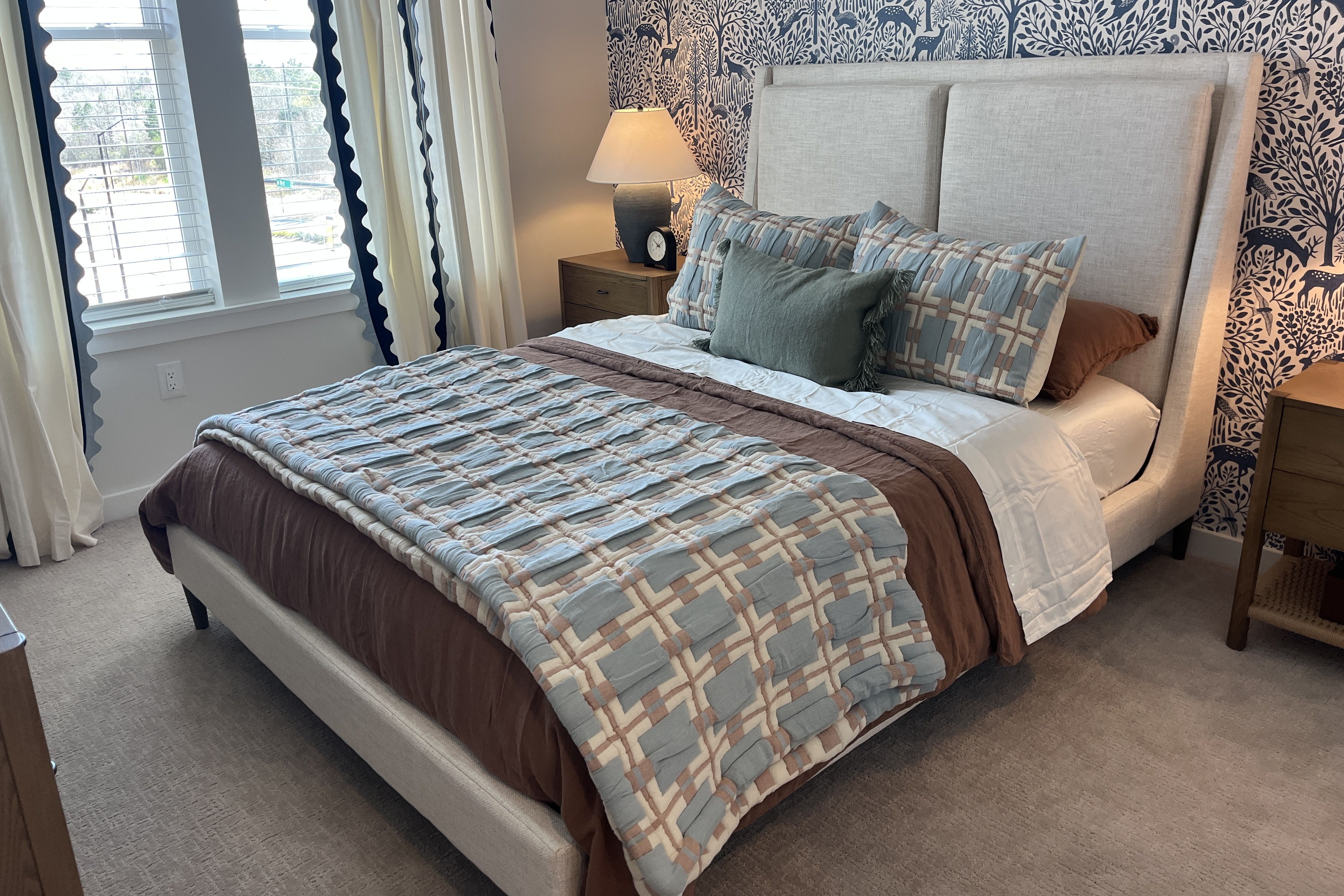 Cozy bedroom with a padded light-gray headboard, blue botanical wallpaper, and a large bed with brown and white linens and a plaid blue-brown quilt. Two wooden nightstands with lamps flank the bed.