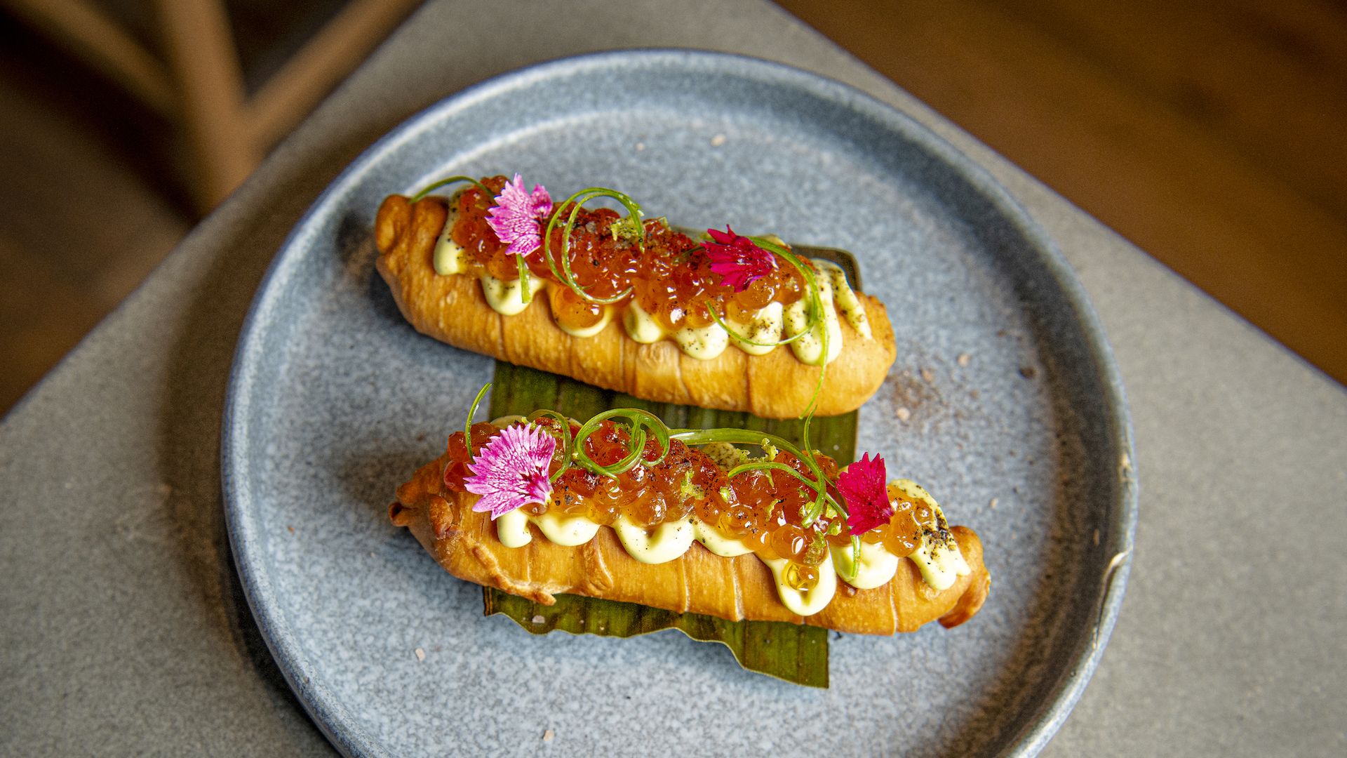 Two golden-brown tequenos pastries on a gray plate with green leaf base, topped with dollops of creamy white sauce, orange fish roe, green herbs, and small pink edible flowers.