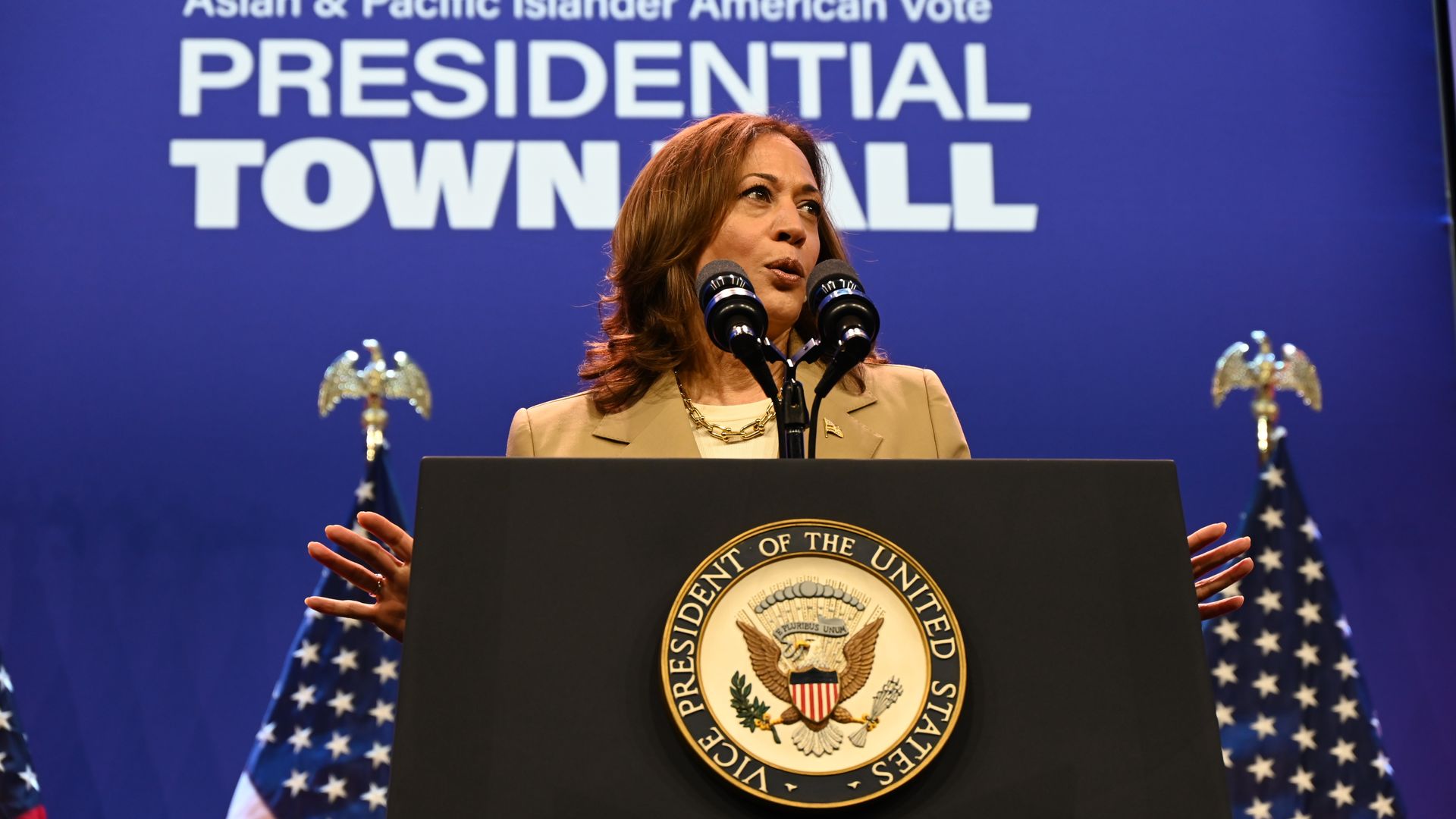 An individual stands at a lectern with a presidential seal affixed to the front. "Asian & Pacific Islander American Vote Presidential Town Hall" on a blue wall and two U.S. flags are in the background