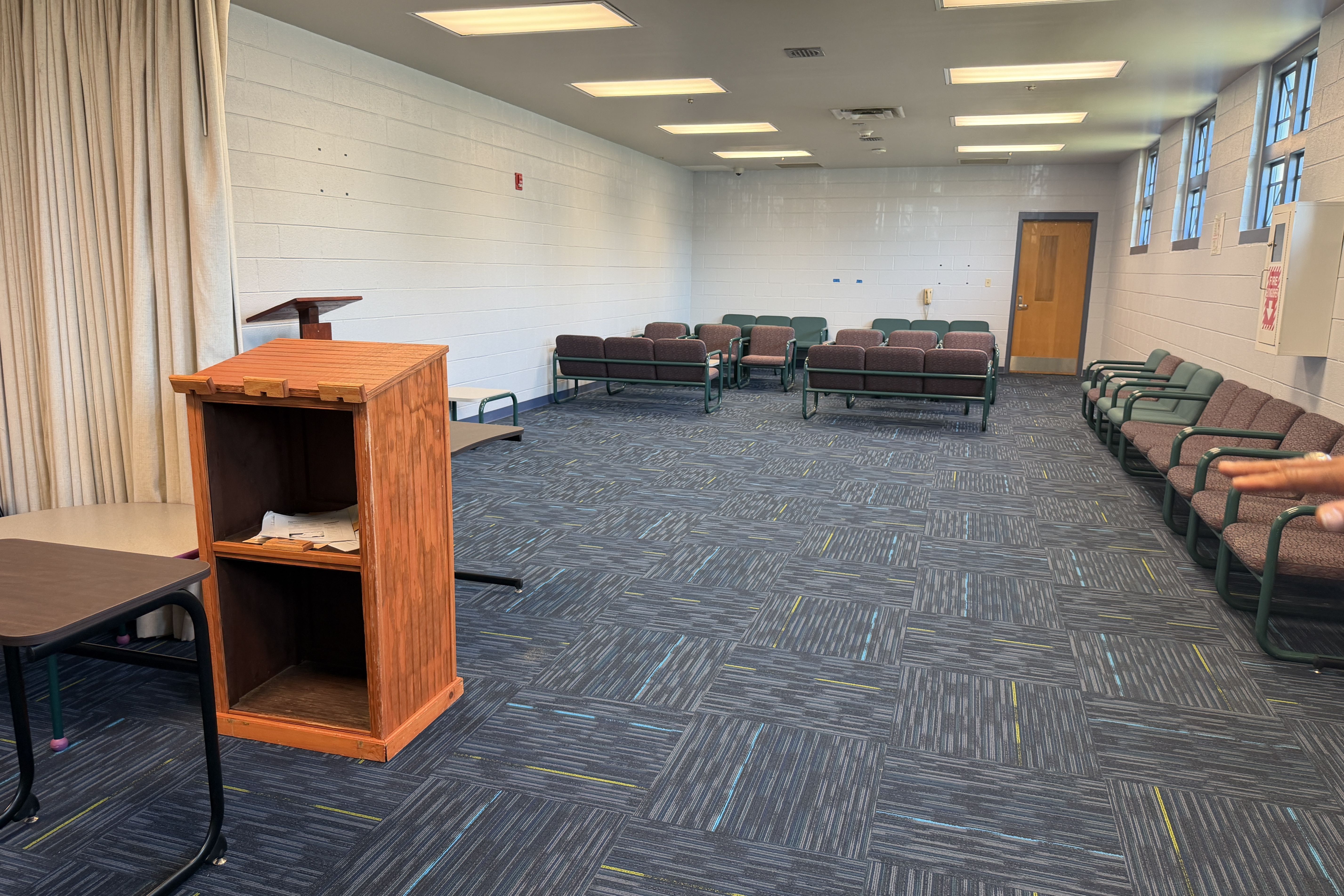Room with a wooden podium on the left, beige curtains, white cinder block walls, rows of green and maroon chairs, blue patterned carpet, bright ceiling lights, and a door with windows on the right.