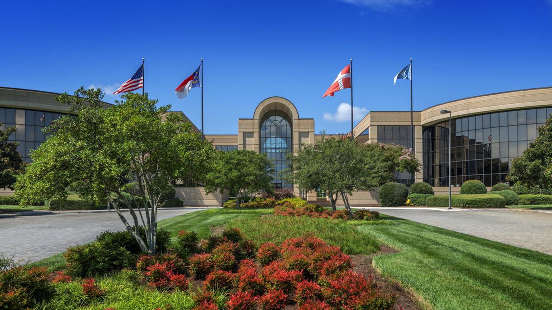Modern beige office building with large windows, surrounded by green trees, red and green shrubs, and four flags including the U.S. flag, under a clear blue sky.