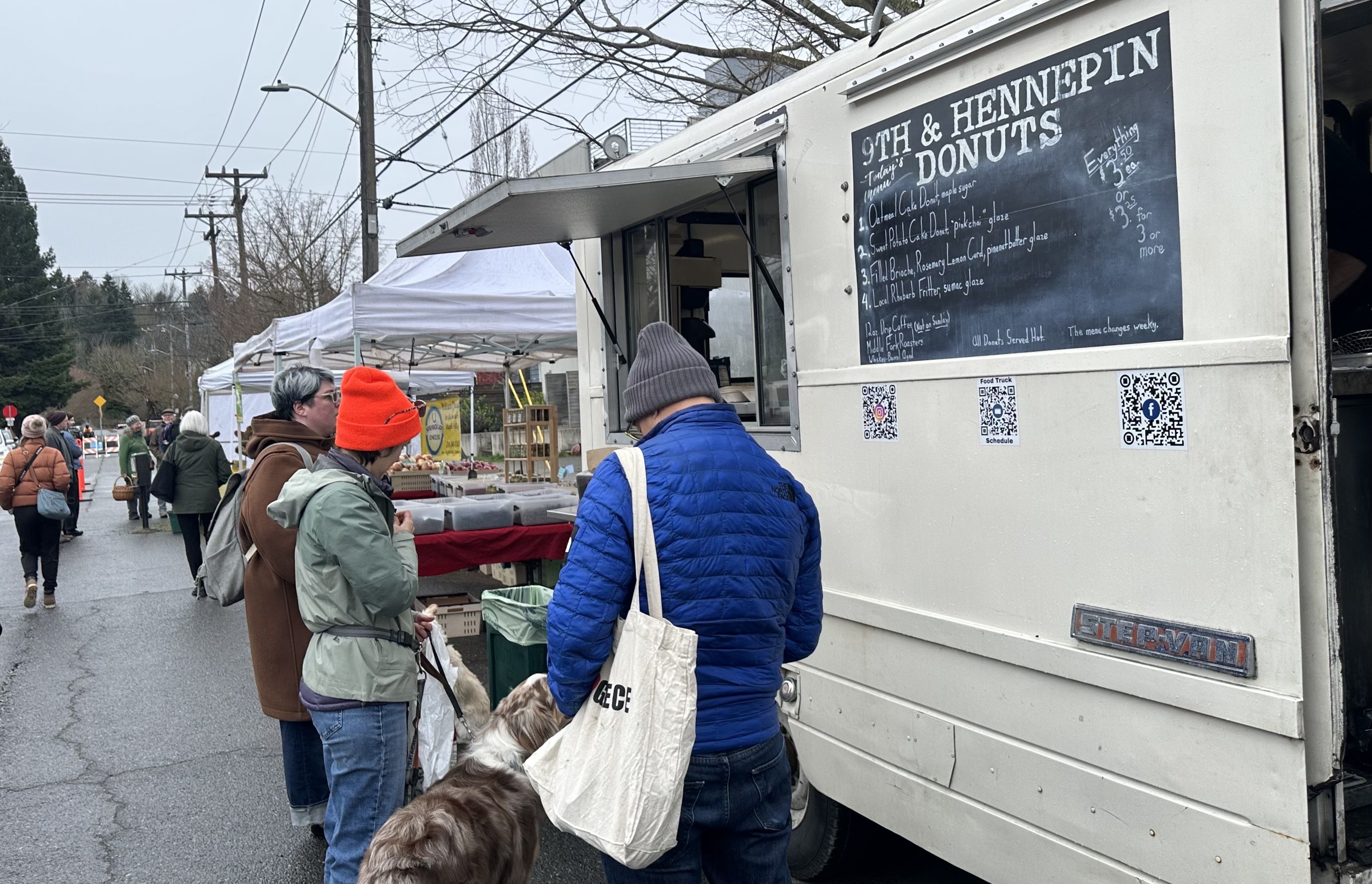 People lined up outside a white truck that says 9th and Hennepin Donuts, with a menu written in chalk. A dog is on a leash waiting, too.
