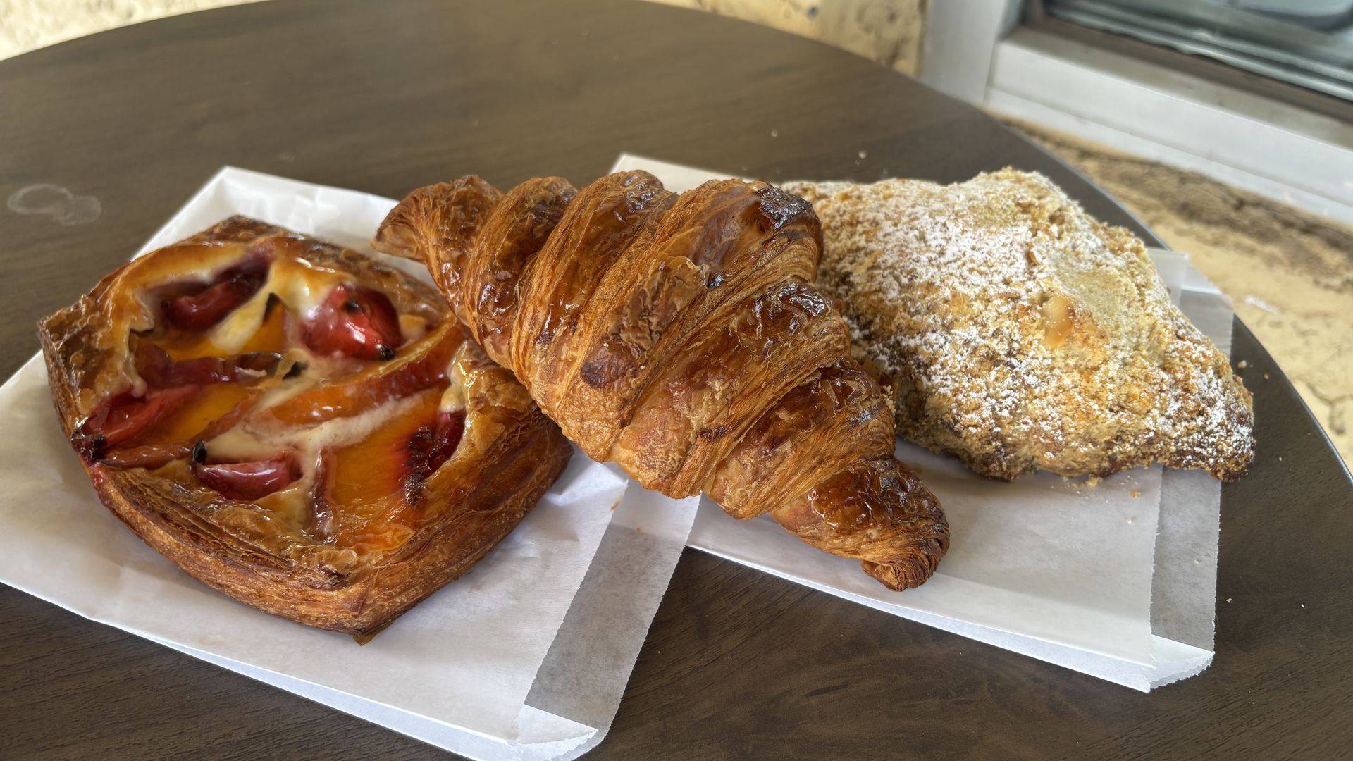 Three pastries on parchment on a dark wooden table: a glossy croissant center, a peppery Danish left, and a sugar-dusted pastry right, with a textured stone wall and window in the background.