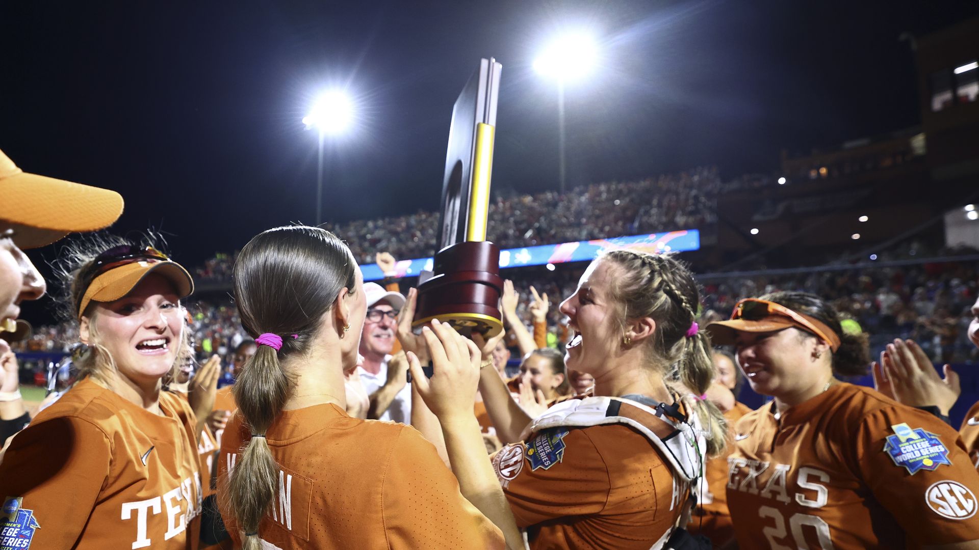 Texas team in orange uniforms celebrate a victory at night, raising a tall trophy amid bright stadium lights, with a large cheering crowd in the stands.