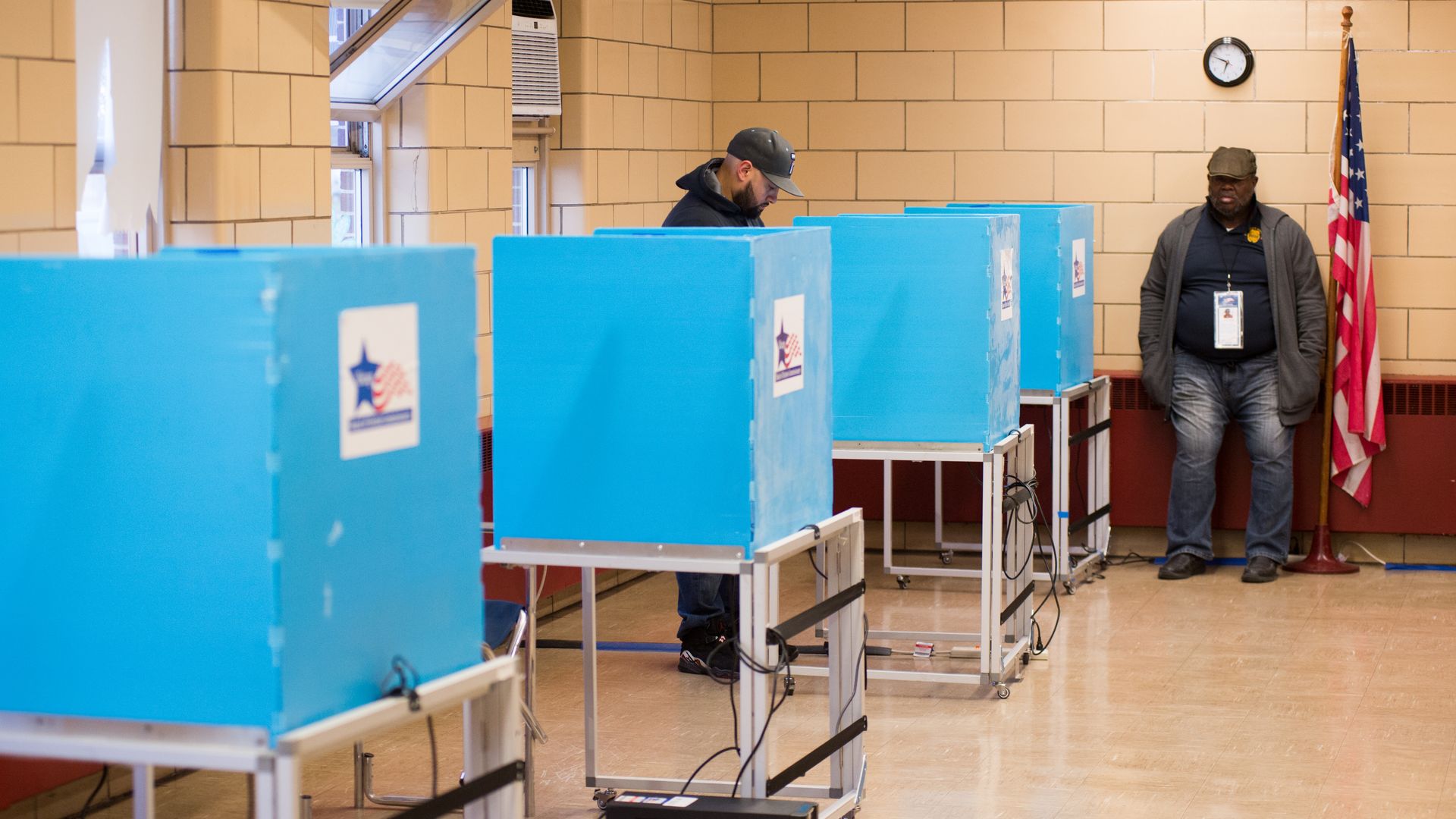 Indoor polling station with beige tiled walls, blue voting booths, a man in a cap and hoodie looking down at a booth, and another man standing by an American flag and a clock.