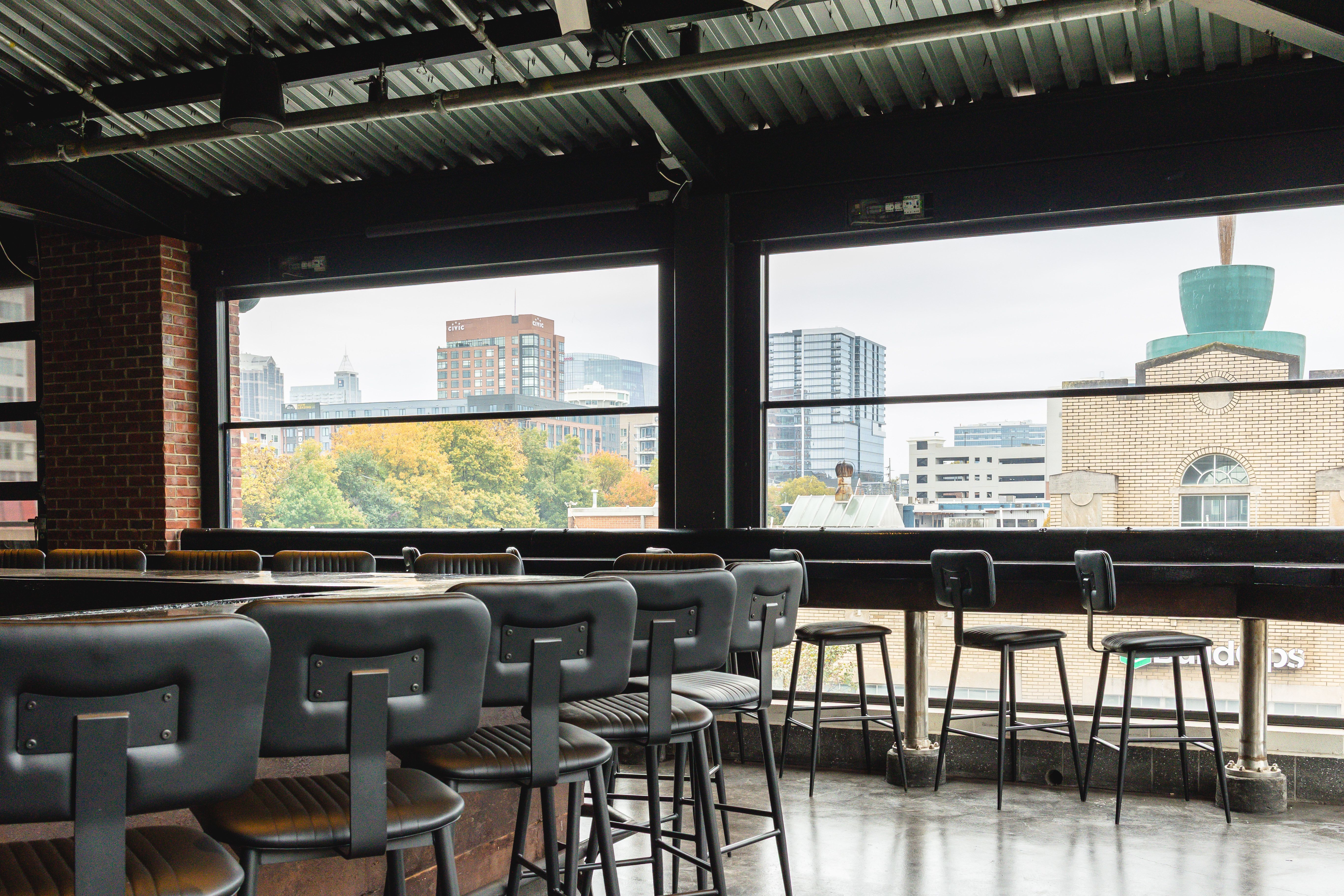 Empty bar seating with black stools and tables inside a building, large windows showing a cityscape with office buildings, trees, and a brick building with a large turquoise cup sculpture on its roof.