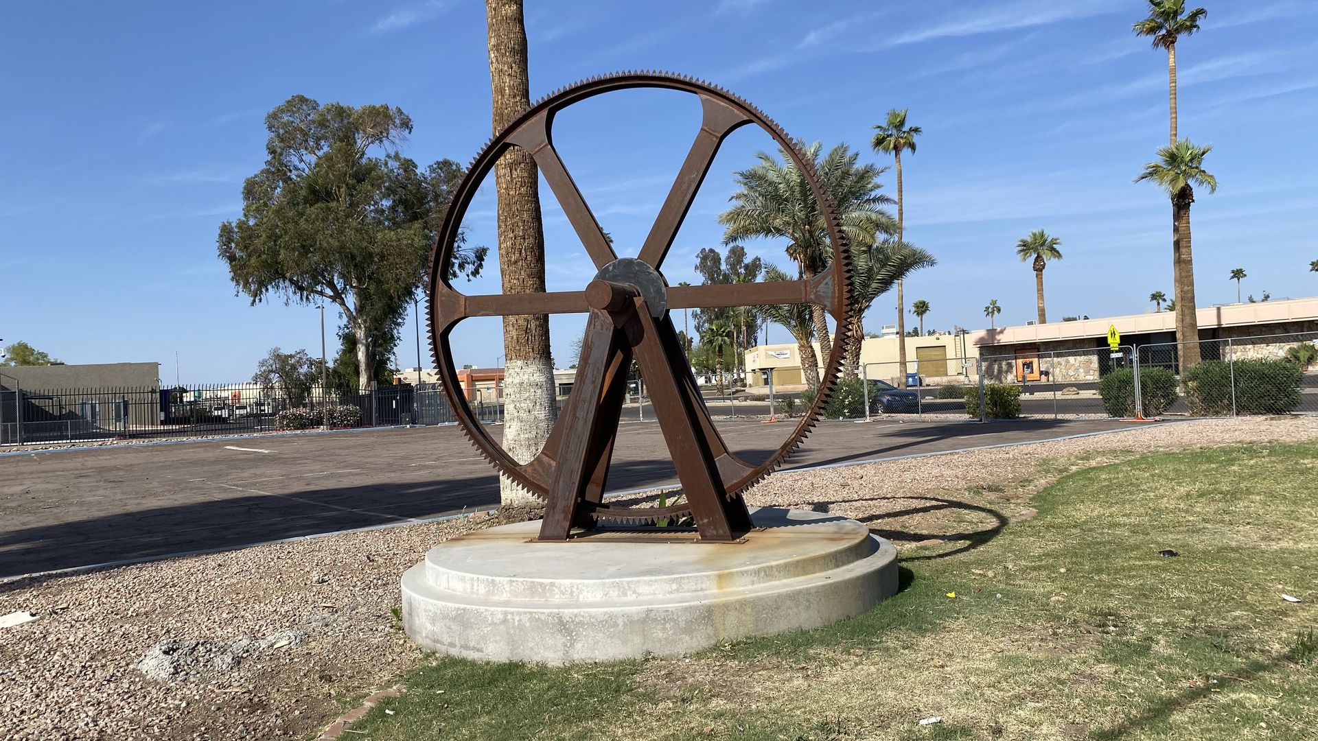 A large rusted metal wheel with six spokes, held in place by an axle, on a concrete pedestal. 