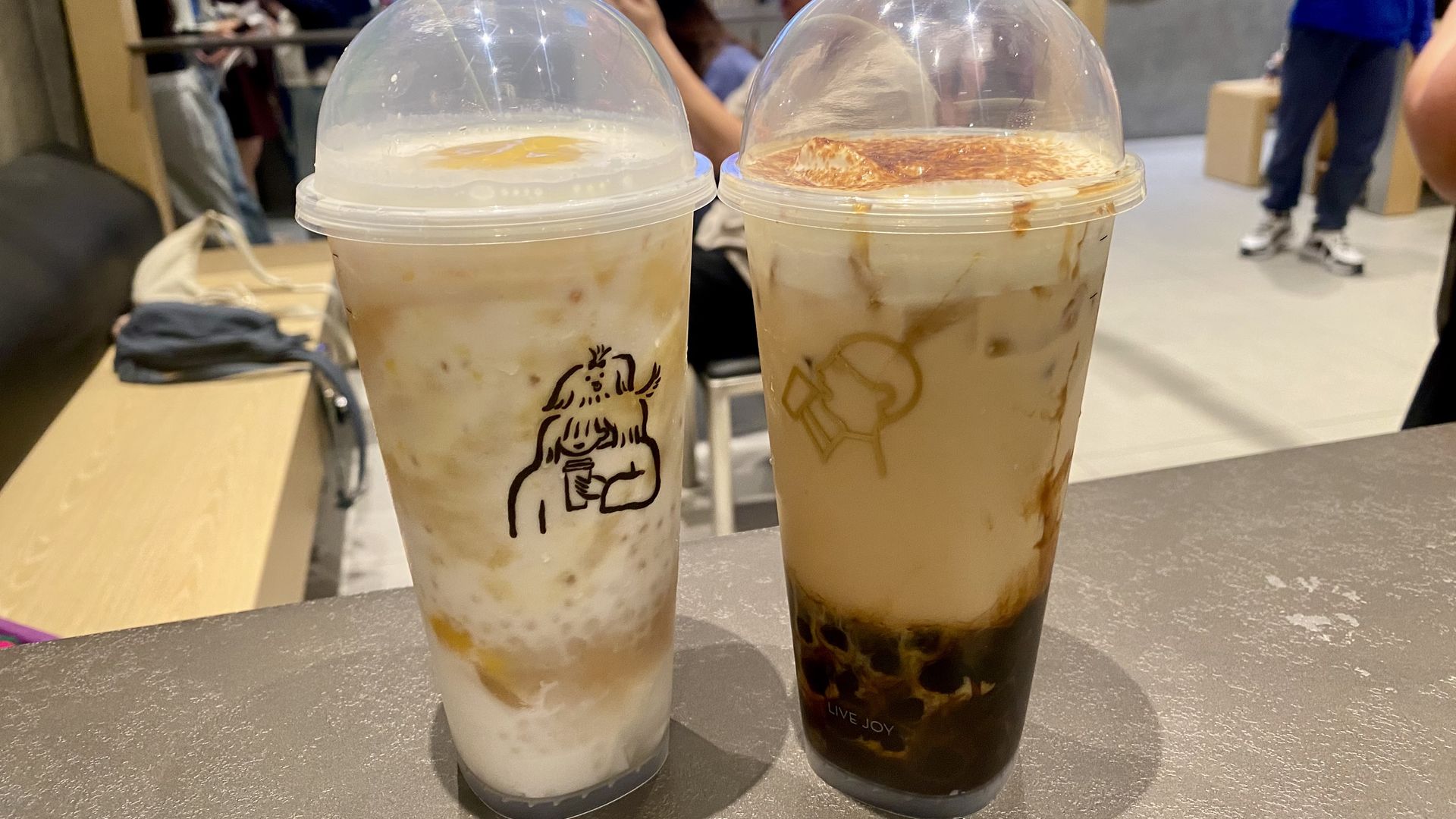 Two bubble tea drinks on a table inside a cafe with people in the background; one drink is white with yellow swirls and a cartoon of a person drinking, the other is beige with brown pearls and foam.
