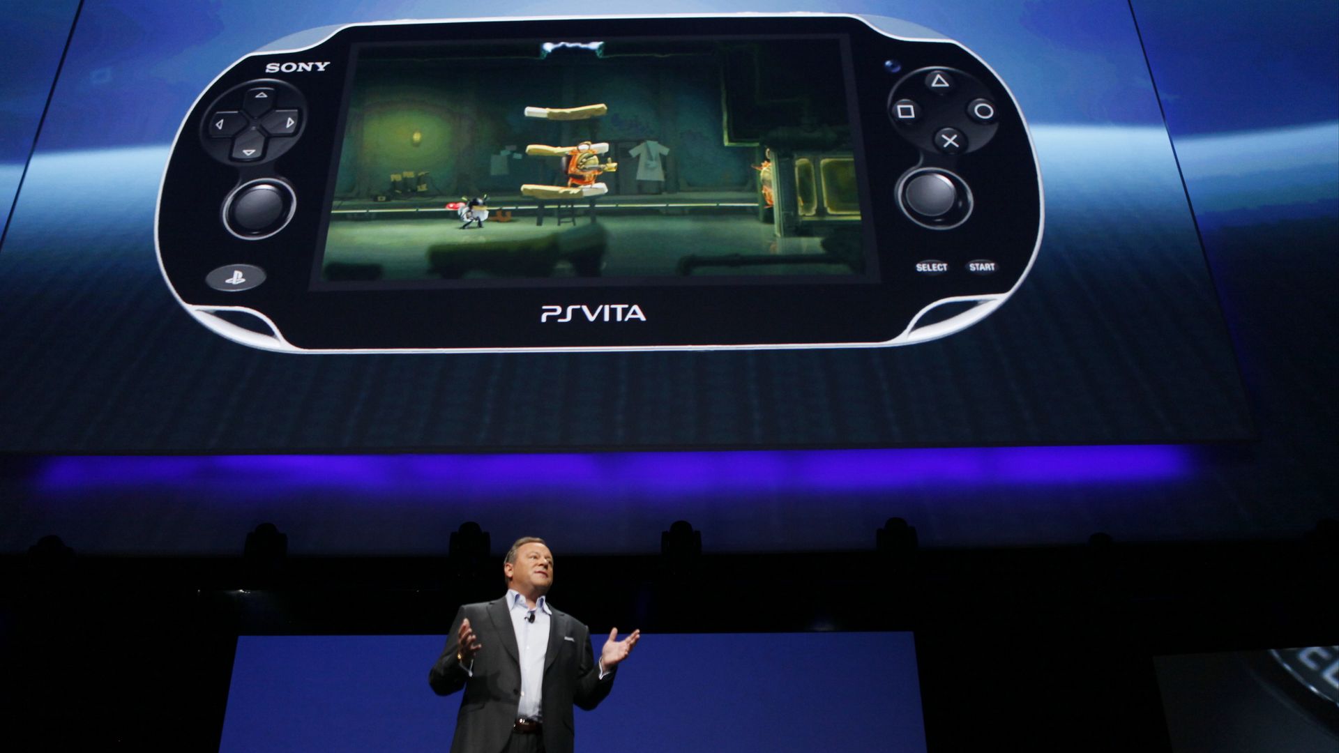 Photo of Jack Tretton in a suit standing on stage below a projection of a large portable gaming device