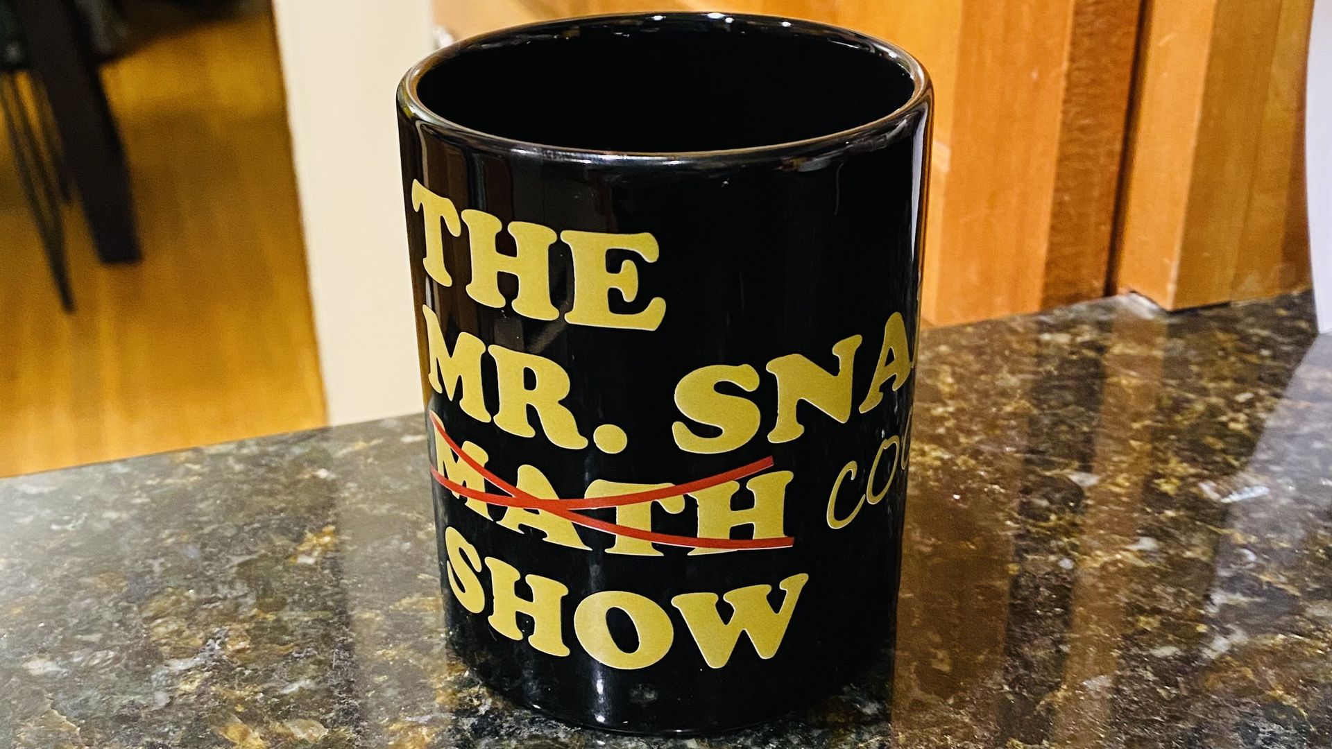 A black mug with yellow letters that reads "The Mr. Snabes Math Show," with "math" crossed out in red and replaced with the word "cooking"
