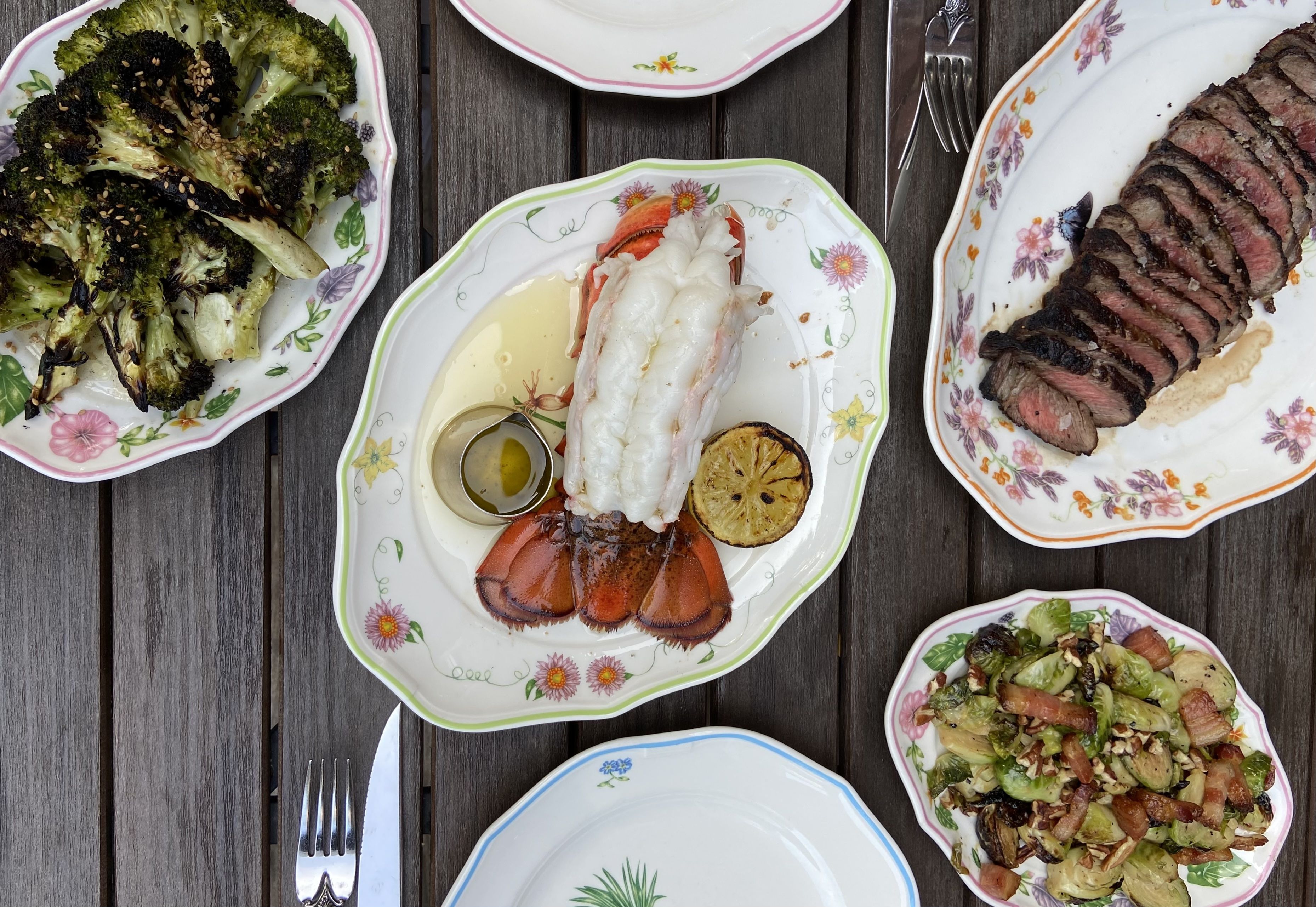 An overhead view of a wooden table set with various dishes. There is a plate of roasted broccoli with sesame seeds, a lobster tail with drawn butter and a slice of grilled lemon, a platter of sliced steak, and a salad featuring Brussels sprouts, bacon, and nuts.