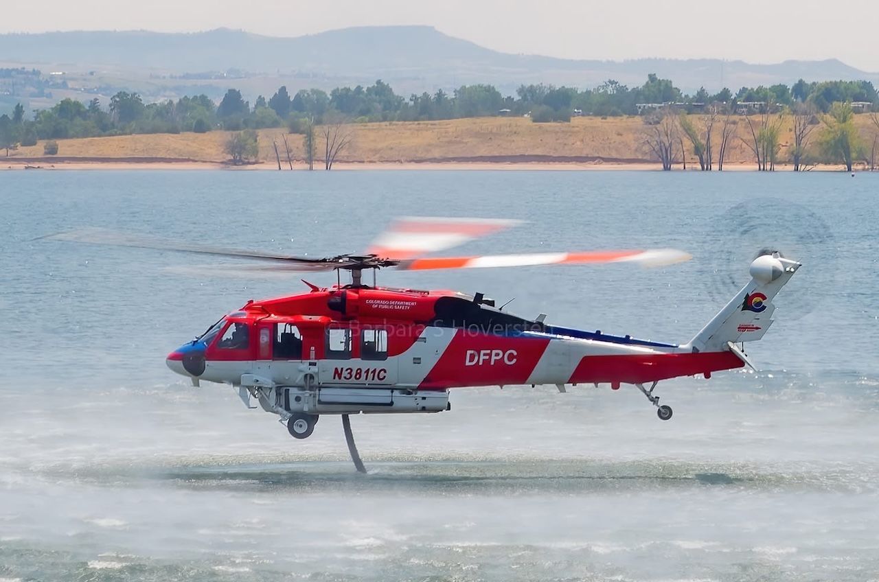 Red and white DFPC rescue helicopter with tail number N3811C hovers over a calm lake, rotor blur as it skims the water; distant shore, trees, and hills in the background.