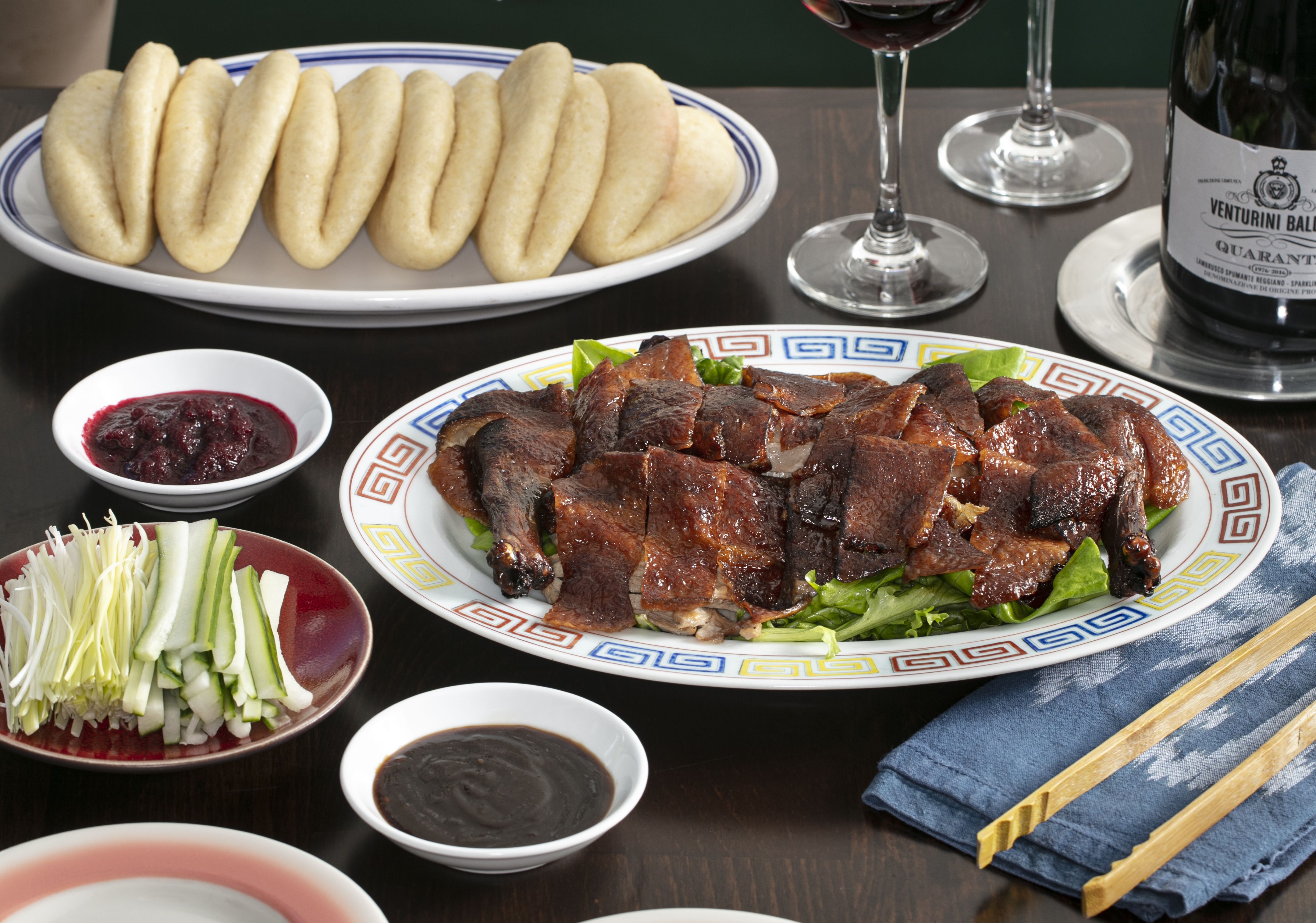 Plate of sliced roasted duck with crispy skin on greens, steamed folded buns, cucumber and scallion strips, dark hoisin sauce, red bean sauce, wine glasses, and chopsticks on a blue napkin.