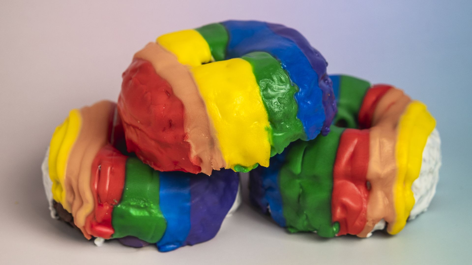 Two donuts topped with thick rainbow frosting in red, orange, yellow, green, blue, and purple stripes, set against a soft pastel gradient background.