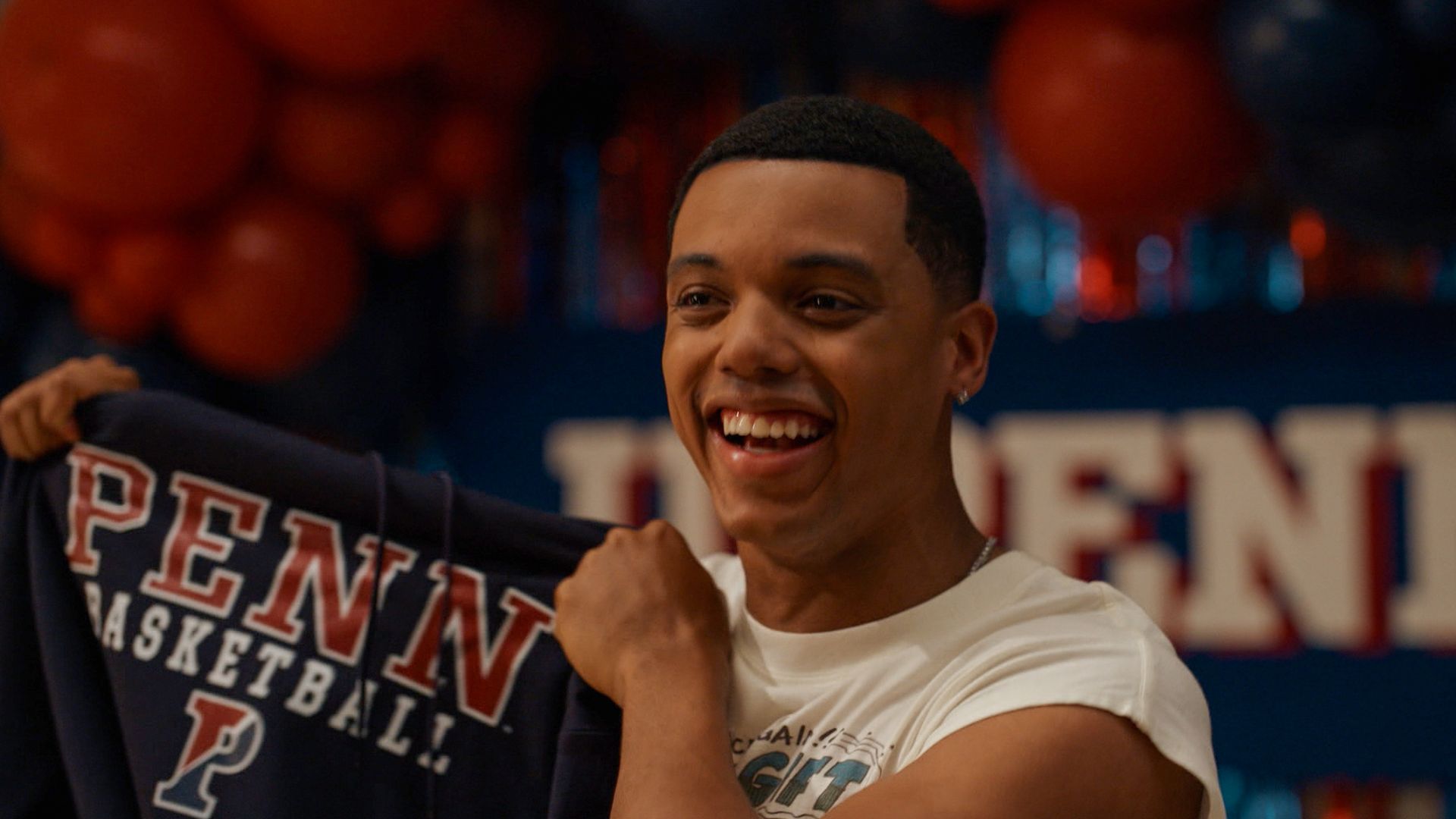 Smiling young man holding a navy blue sweatshirt with red and white lettering that reads "PENN BASKETBALL" against a background with red balloons and blurred similar lettering.