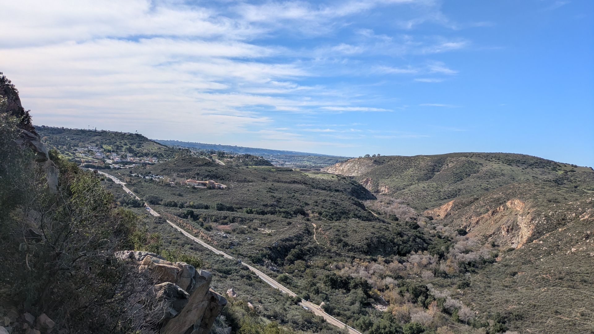 Wide valley view with rolling brown and green hills, a winding road through the slope, rocky foreground on the left, sparse trees, and a distant town under a bright blue sky with wispy clouds.