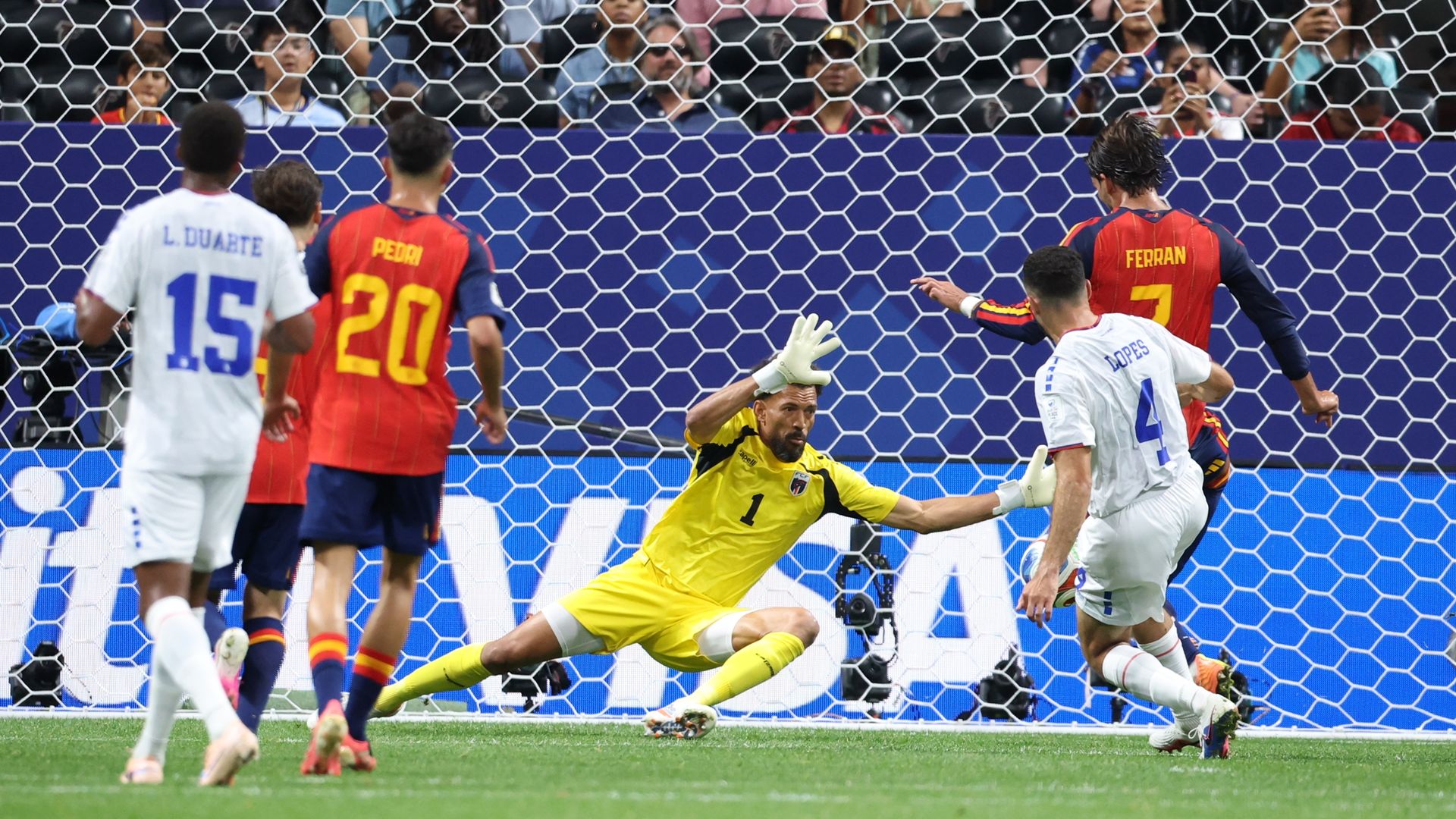 A goalkeeper wearing a yellow kit blocks a shot attempt by a player wearing red and blue with yellow lettering. The goalie's teammates wearing white watch the action.