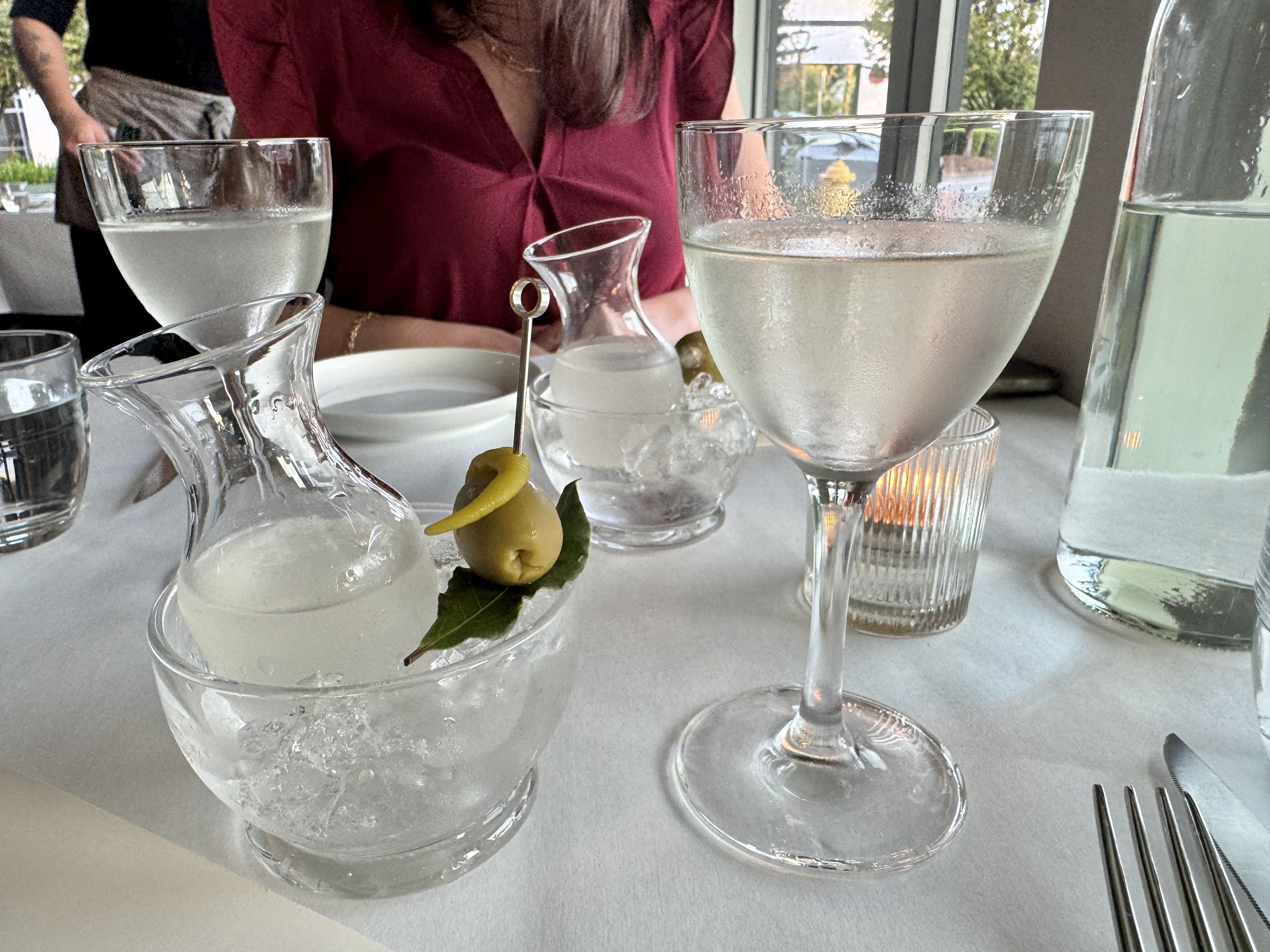 A table set for drinks with clear martini glasses, a small carafe on ice with an olive and pepper garnish, a candle, water glass, and a person in a burgundy shirt in the background.