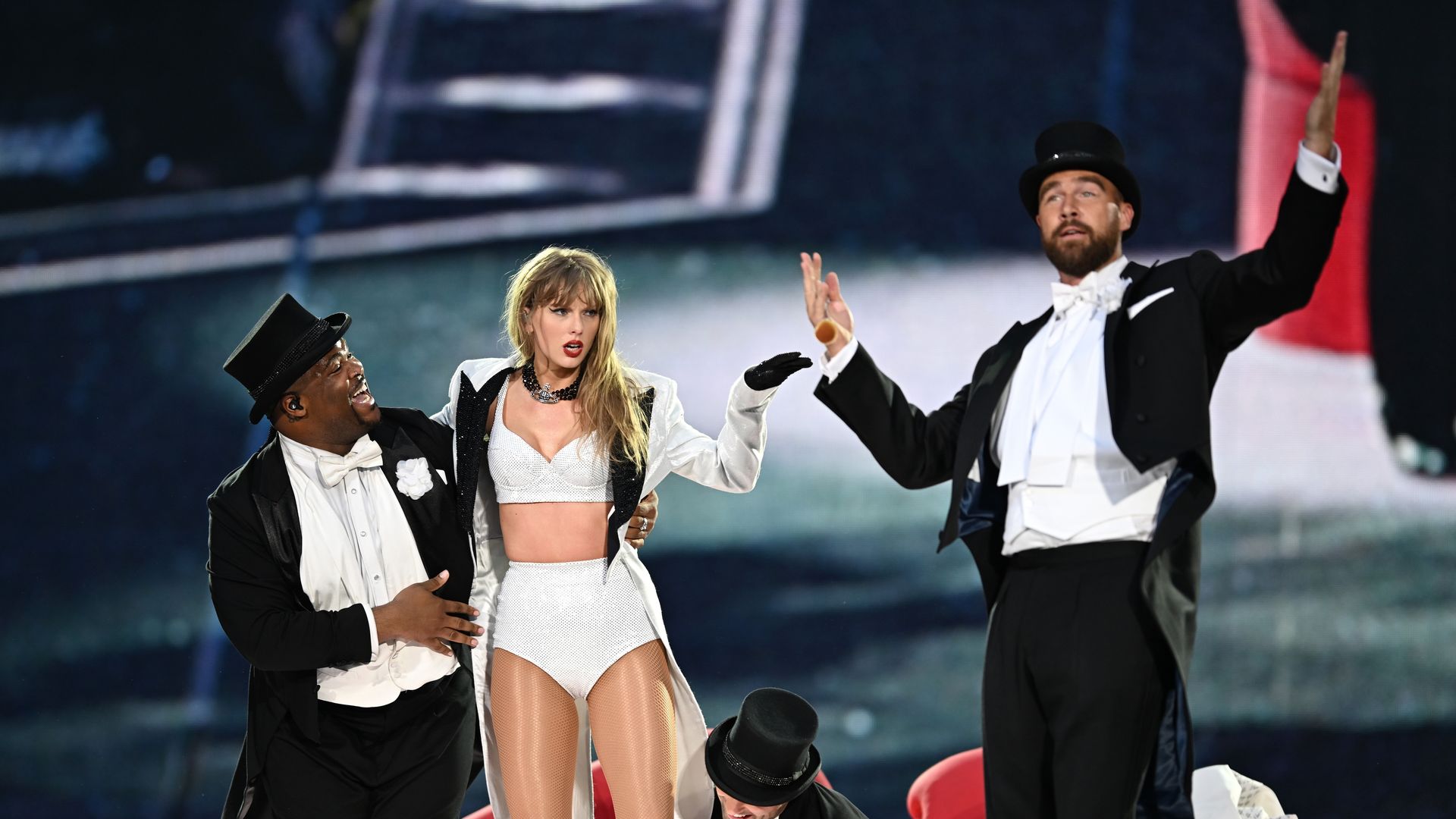 A dancer, Taylor Swift and Travis Kelce in black tuxedos and top hats performing with a woman in white sparkly outfit and fishnet stockings on stage, dramatic expressions and gestures under bright lighting.