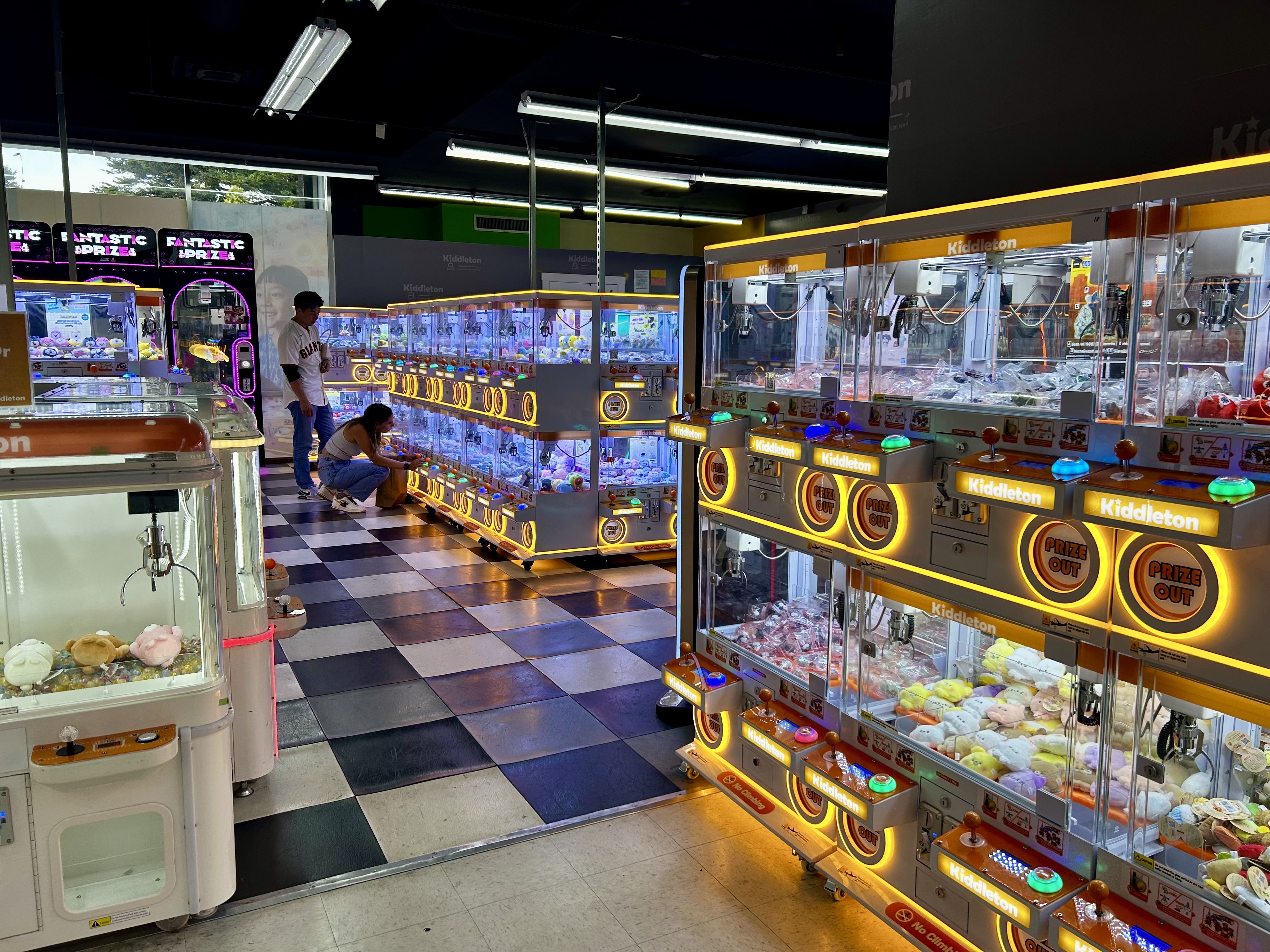 Indoor arcade with rows of brightly lit claw machines labeled "Kiddleton" filled with plush toys. Two people play a machine on a black-and-white checkered floor under fluorescent lights.