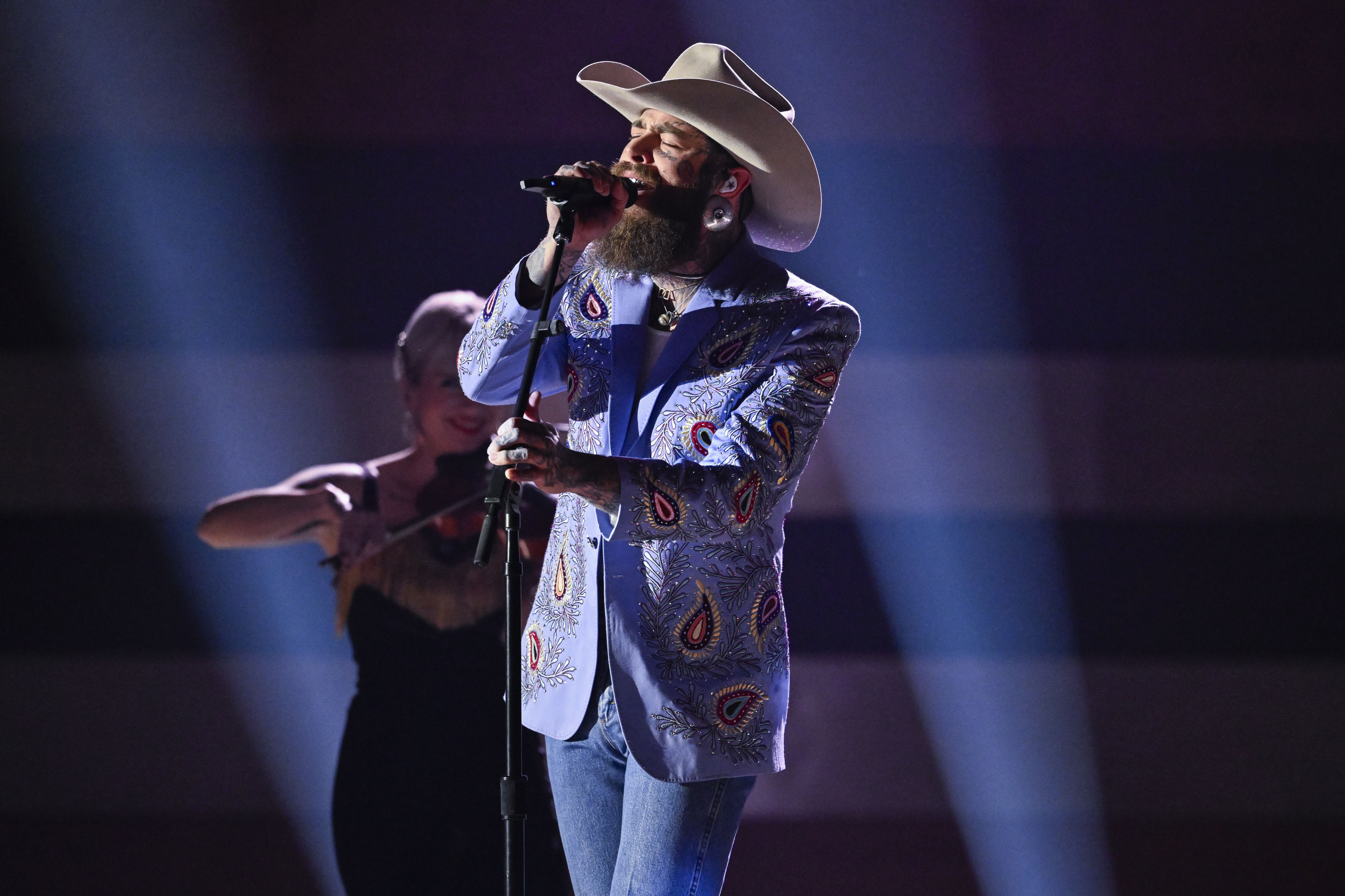 Post Malone, wearing an embroidered suit and a cowboy hat, sings into a microphone.
