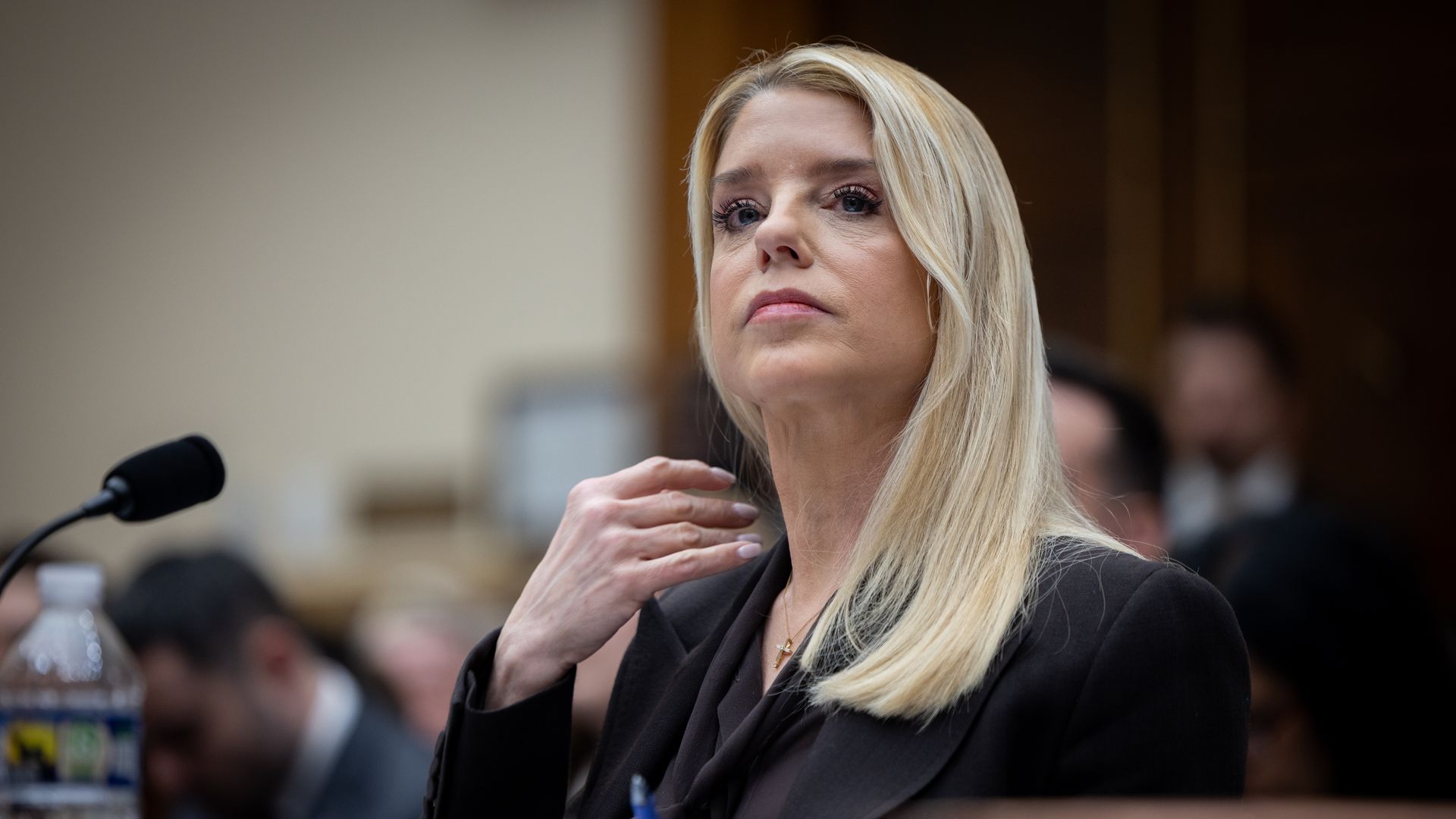 Một người phụ nữ tóc vàng mặc bộ vest đen đang chỉnh tóc trong khi cầm bút, ngồi tại bàn làm việc với micro và chai nước bên cạnh, trong một khung cảnh trang trọng với những người ở phía sau bị làm mờ.