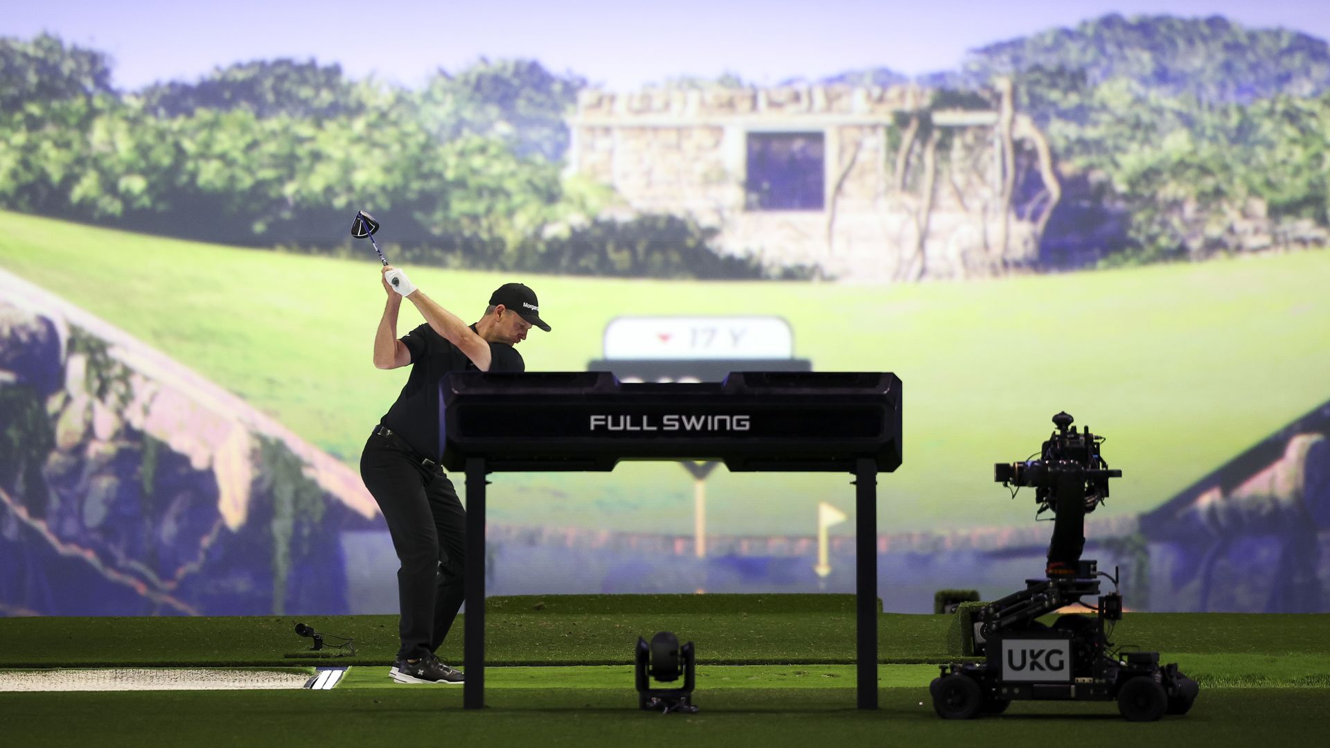 A golfer in black with a cap is mid-swing on a stage with a FULL SWING stand; a camera rig sits to the right on a green field with a large scenic backdrop.