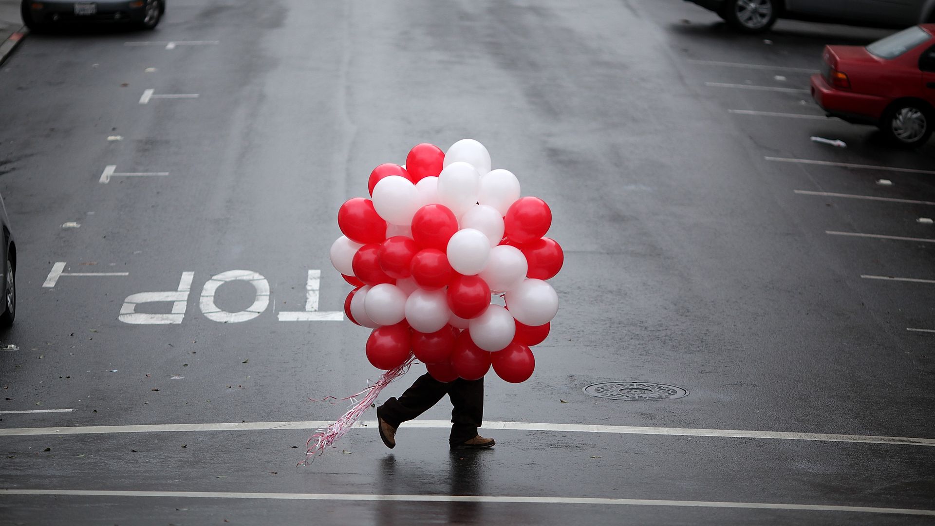 Obscured person carrying large bunch of red, white and pink balloons across street. 