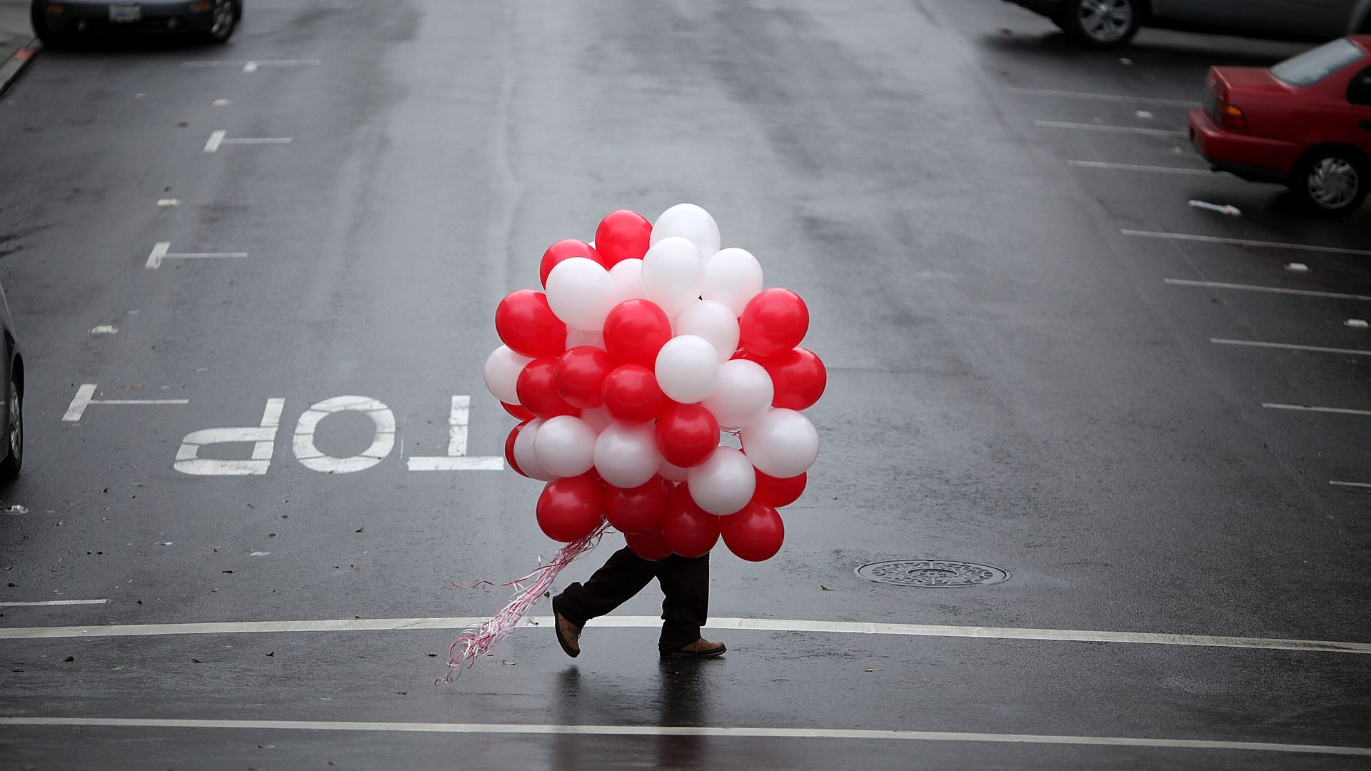 Obscured person carrying large bunch of red, white and pink balloons across street.