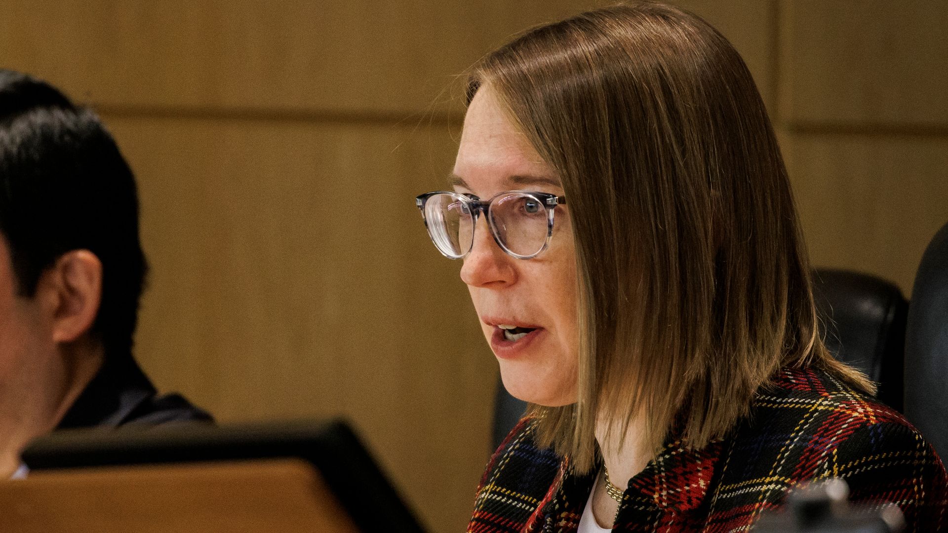 A woman in a plaid suit and glasses, speaking in an official-looking, heavily wood-paneled room.