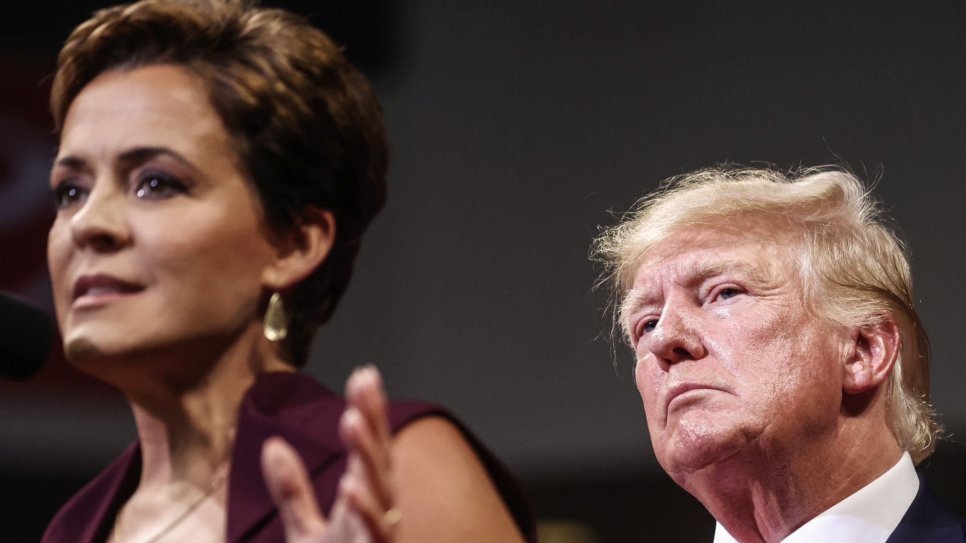 Trump official Kari Lake, who has short, dark hair, dark eyes and a maroon top, gestures with her left hand, as President Trump, wearing a navy jacket, white shirt and red tie, peers over her shoulder.