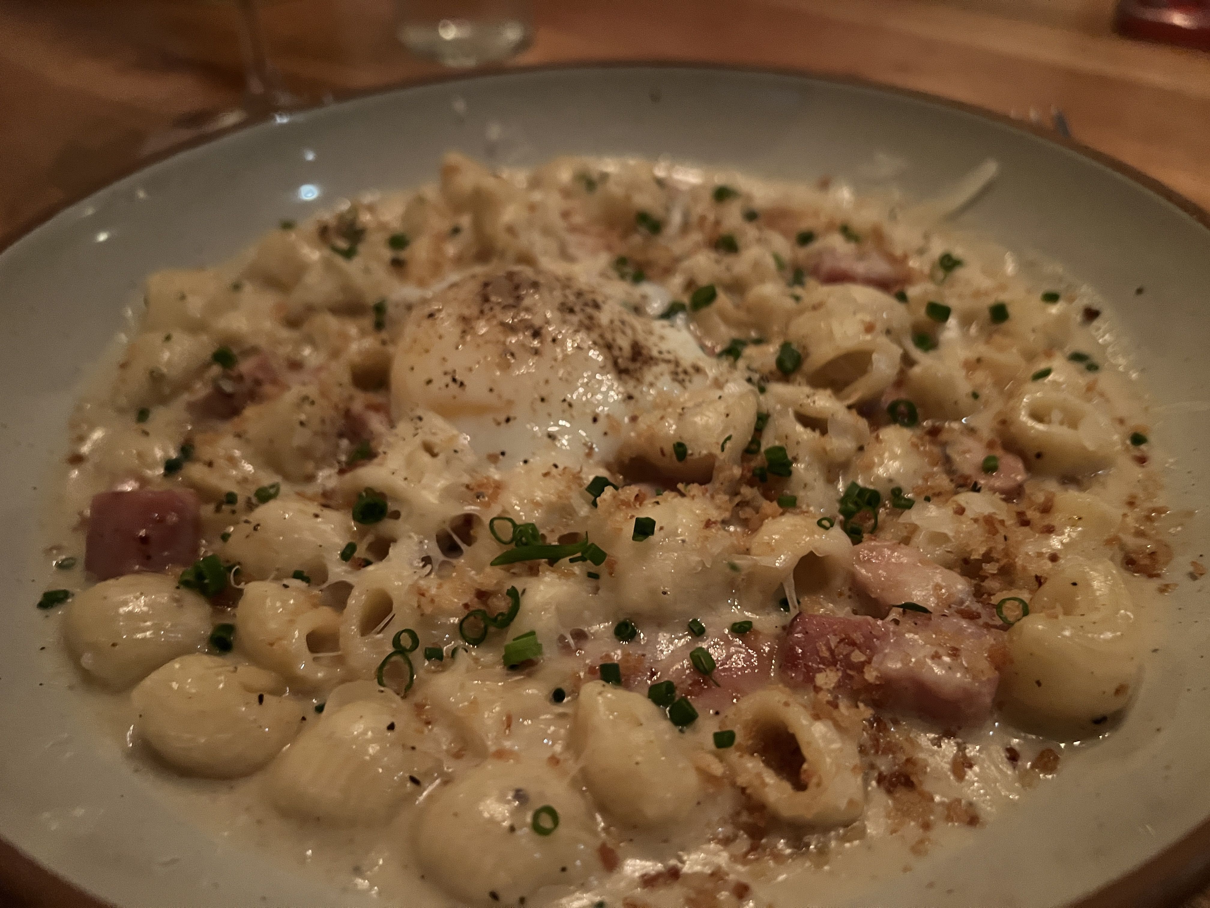 A plate of creamy pasta featuring small shell-shaped pasta with chunks of ham, topped with a runny poached egg. The dish is garnished with chopped chives and a sprinkle of black pepper, creating a rich and indulgent appearance.