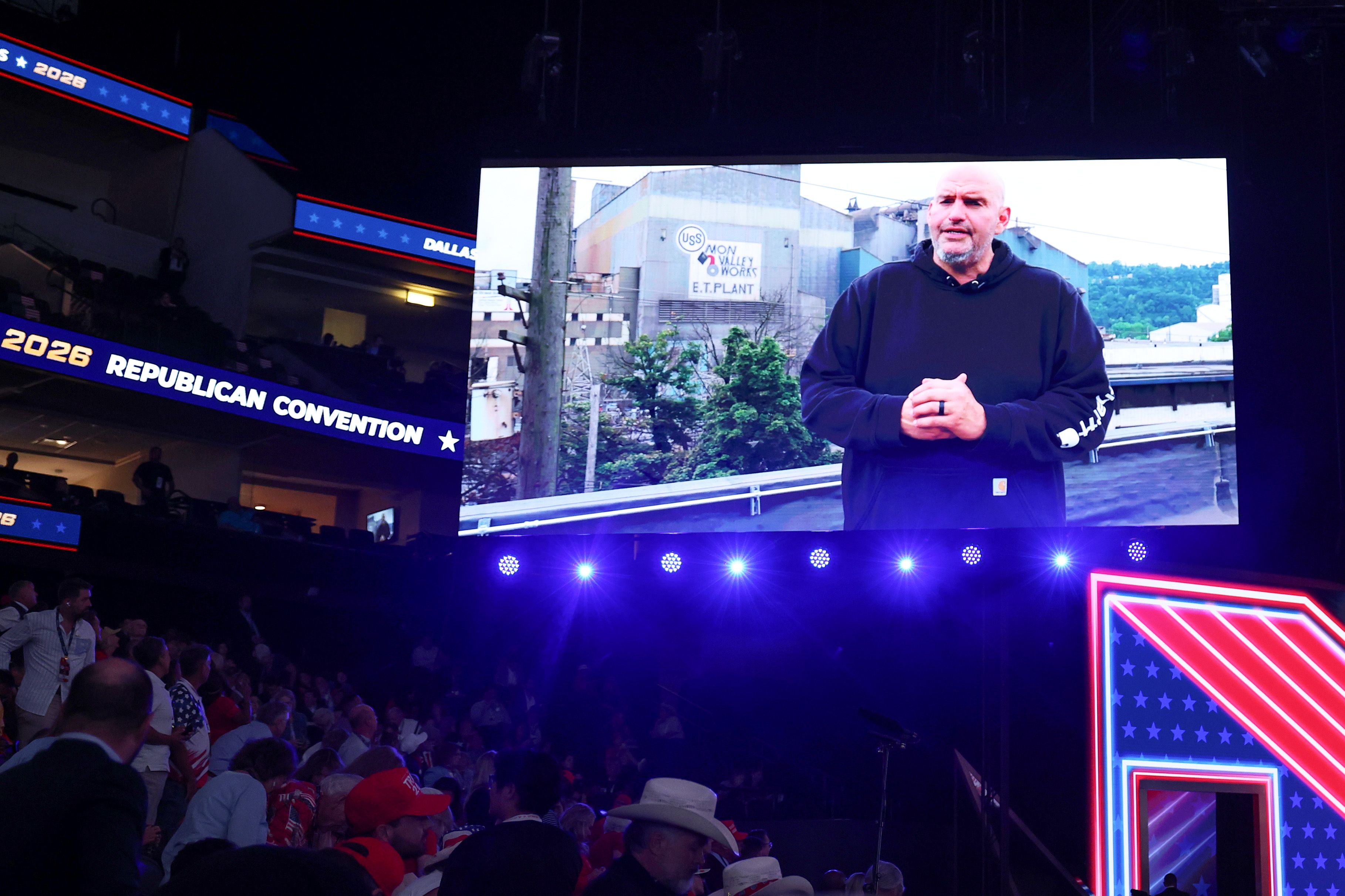 Large stage screen shows Fetterman in a dark hoodie outdoors near an industrial plant; blue stage lights, seated audience, and the "Republican Convention" banner visible.