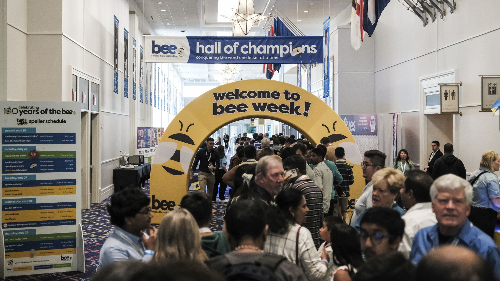 Busy convention hall for a spelling bee; a blue banner reads "bee hall of champions" and a large yellow arch says "welcome to bee week!"; flags line the right wall as crowds fill the corridor.