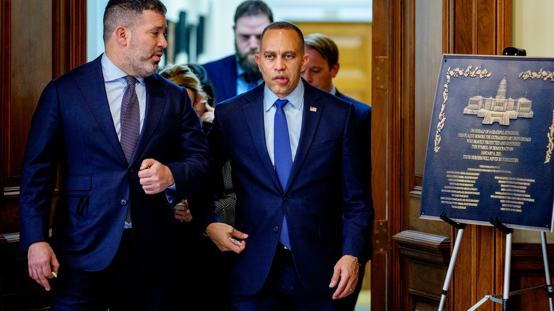 Two men in dark suits and ties walk down a hallway with wooden doors. A plaque on a stand nearby honors those who "defended democracy" on January 6, 2021.