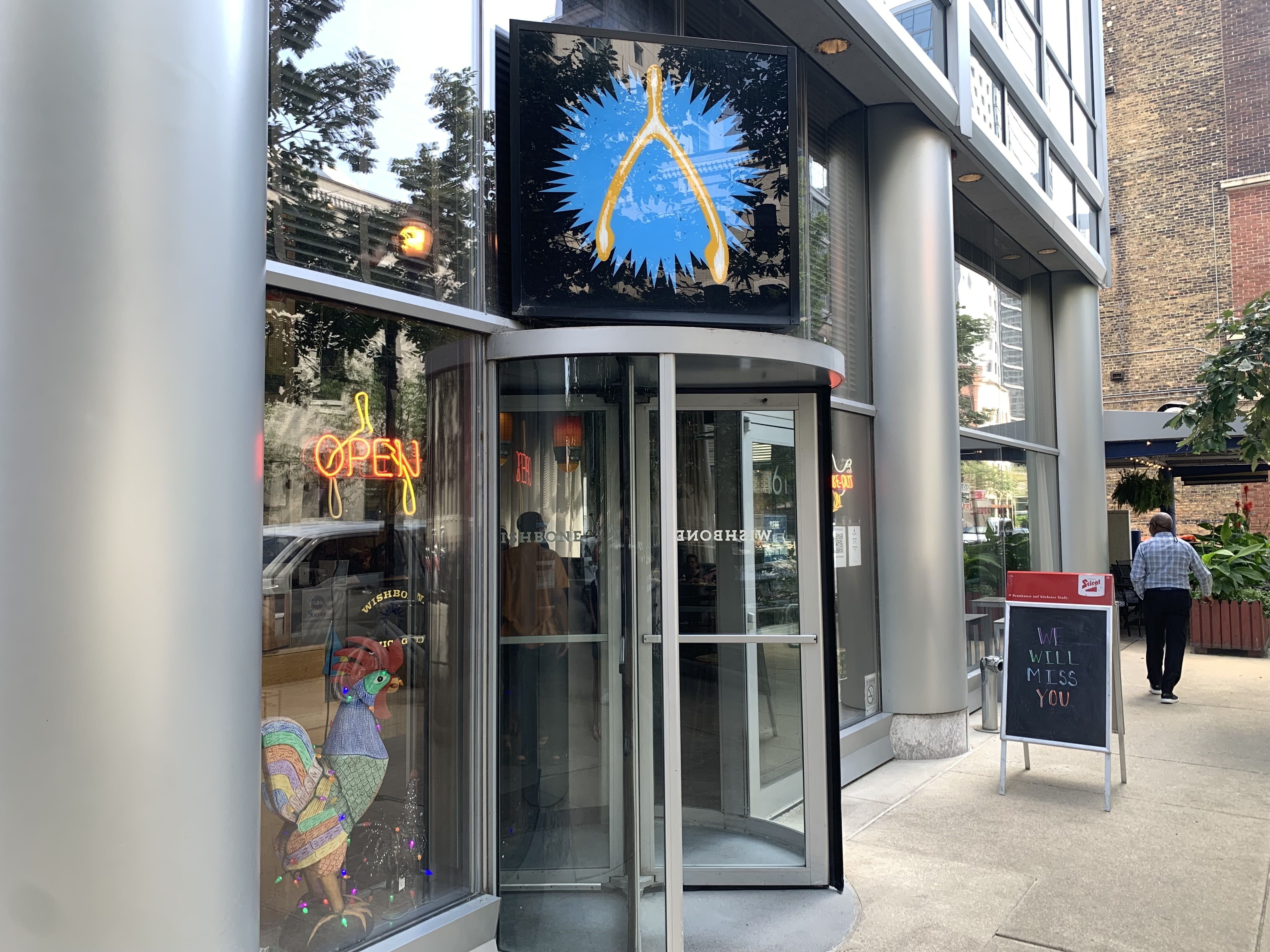 Storefront with a black-framed sign showing a blue burst and yellow umbrella shape. Glass windows reflect trees; neon orange "OPEN" sign and a chalkboard outside; a man walks by on the right.