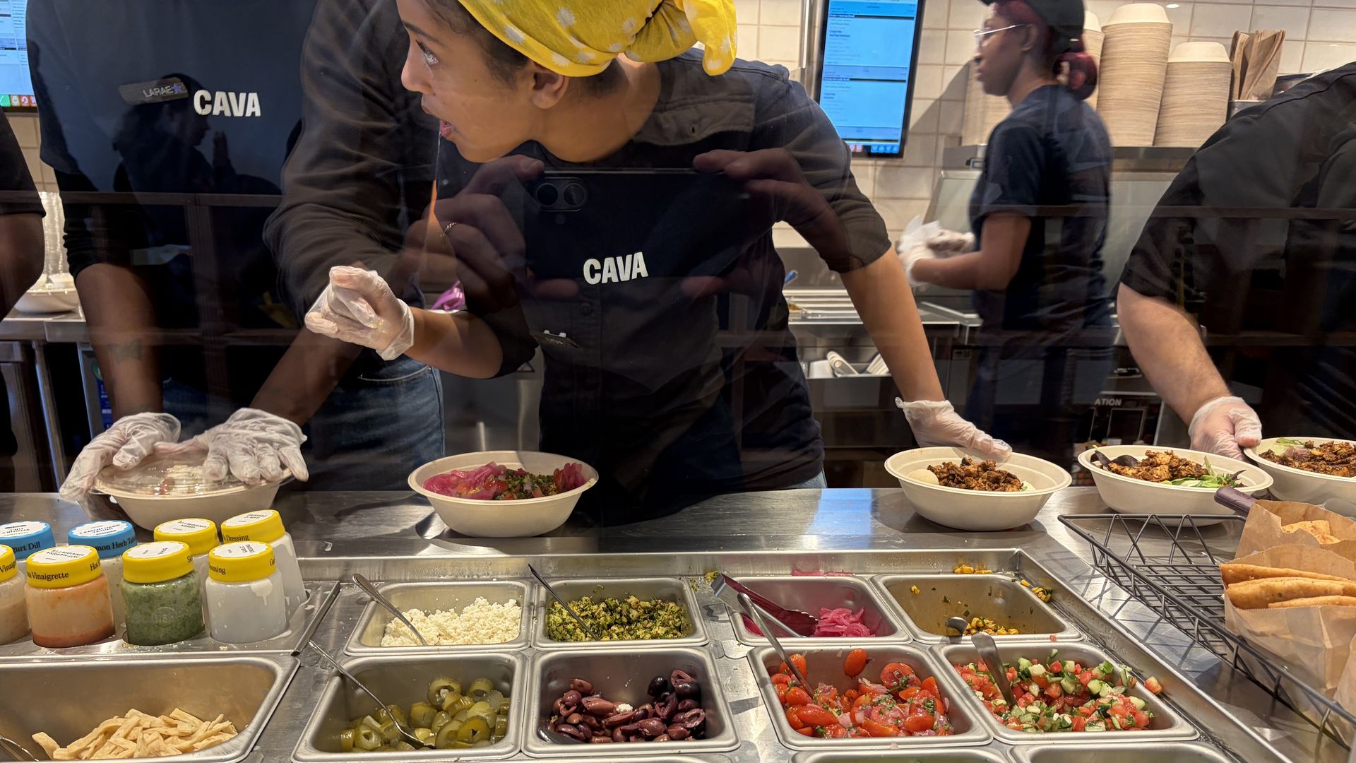 Two CAVA workers in black uniforms behind a glass salad bar, one wearing a yellow headscarf, assembling bowls of greens, olives, tomatoes, peppers, and falafel as sauces line the counter.