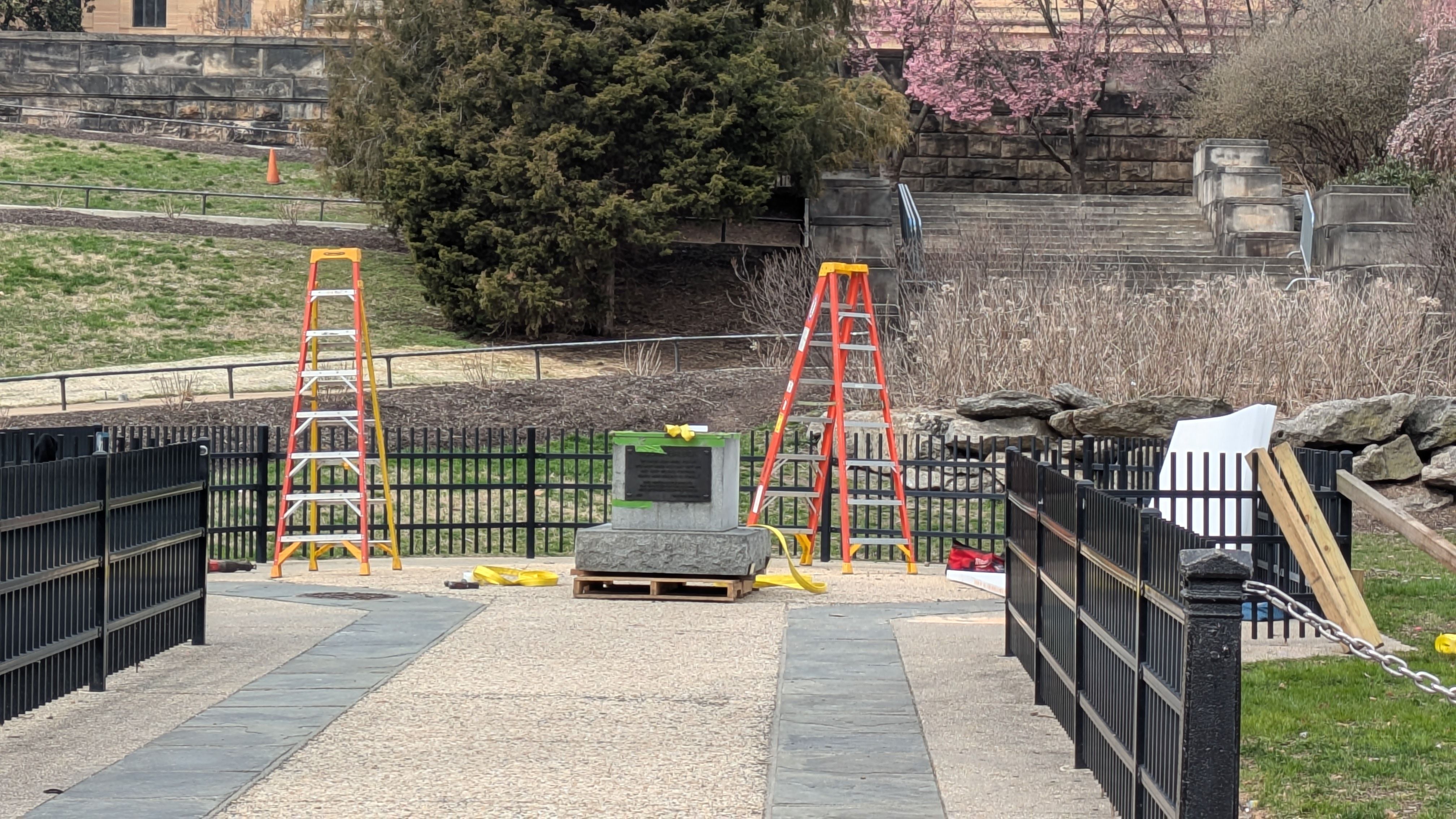 Park scene with a stone pedestal monument on a pallet, two tall ladders (yellow and orange), yellow tape and tools nearby; black fences line a path, with blooming trees and stone steps in back.