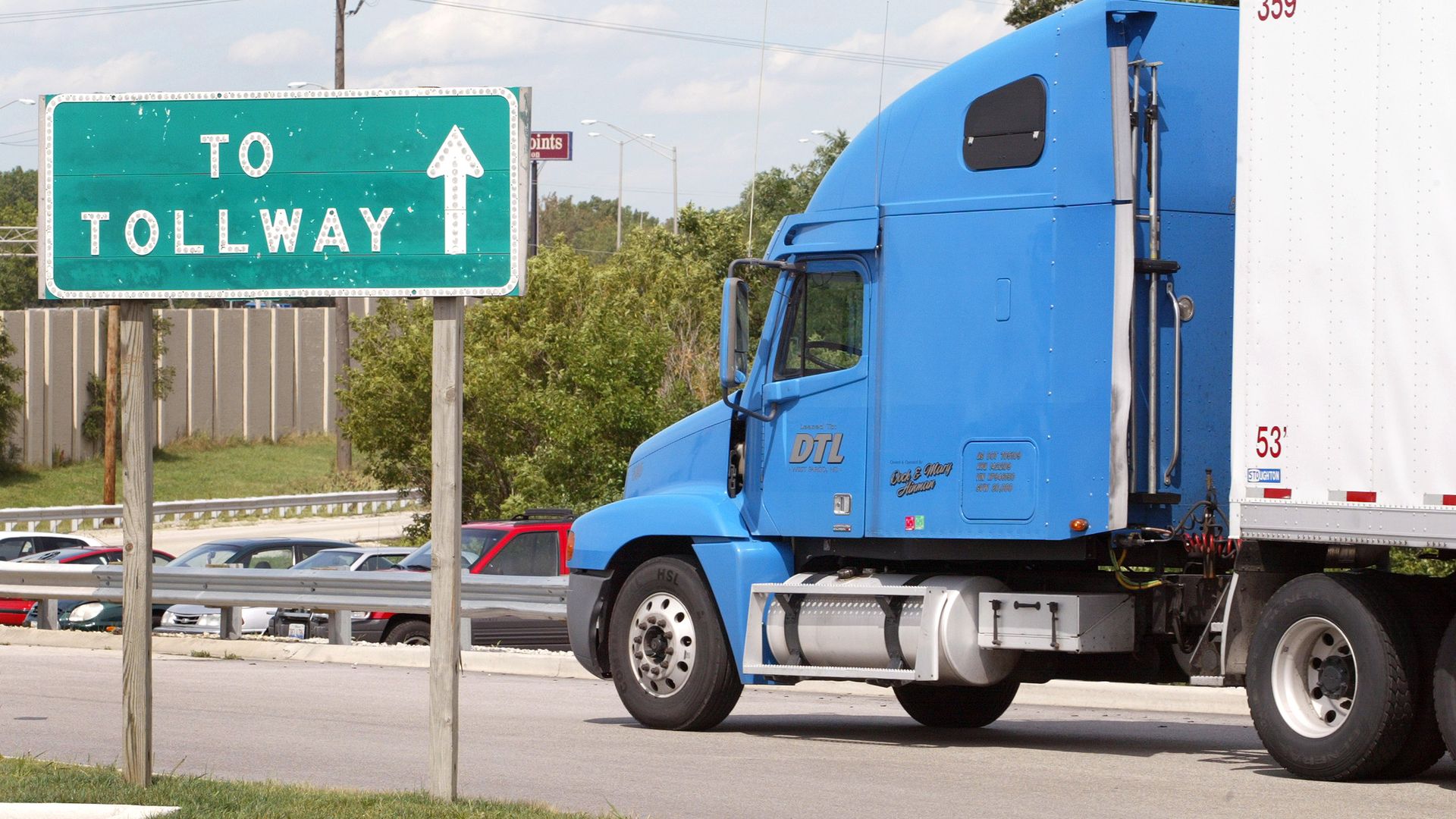 A blue semi-truck with a white trailer on a highway beside a green "To Tollway" sign with an upward arrow; trees, a fence, and parked cars in the background.