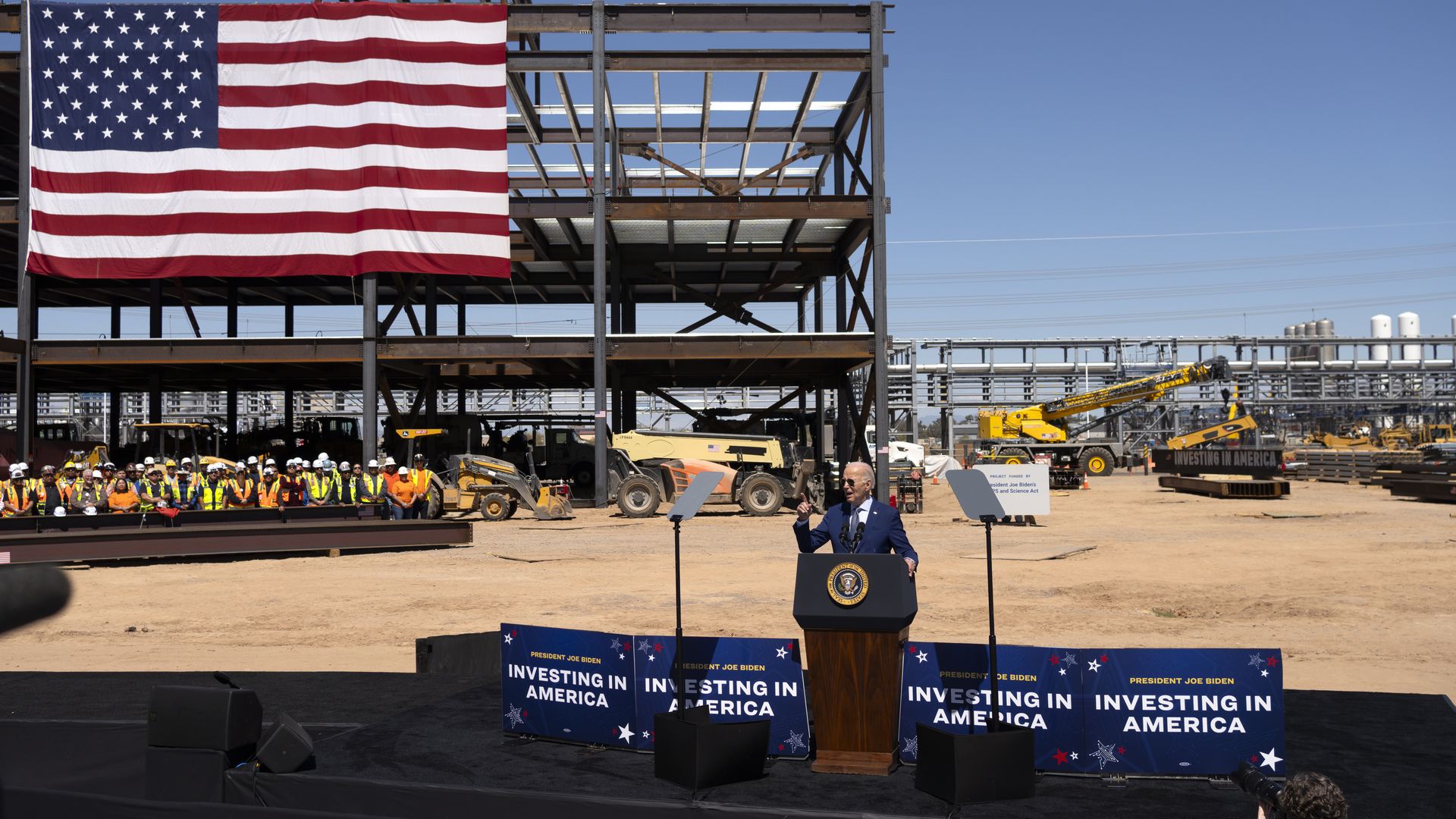 President Biden standing on a stage with a worksite in the background and a large American flag.