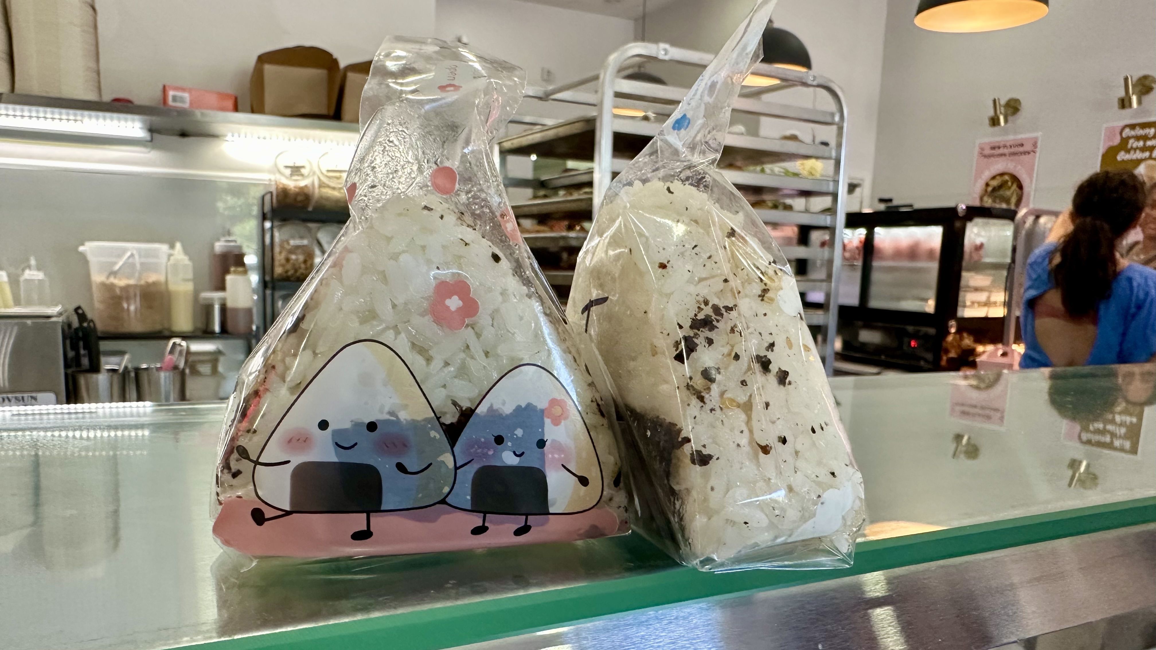Two clear plastic bags with cute pastel mountain graphics and smiling faces sit on a glass bakery counter; background shows shelves, a worker in blue, and warm hanging lights.