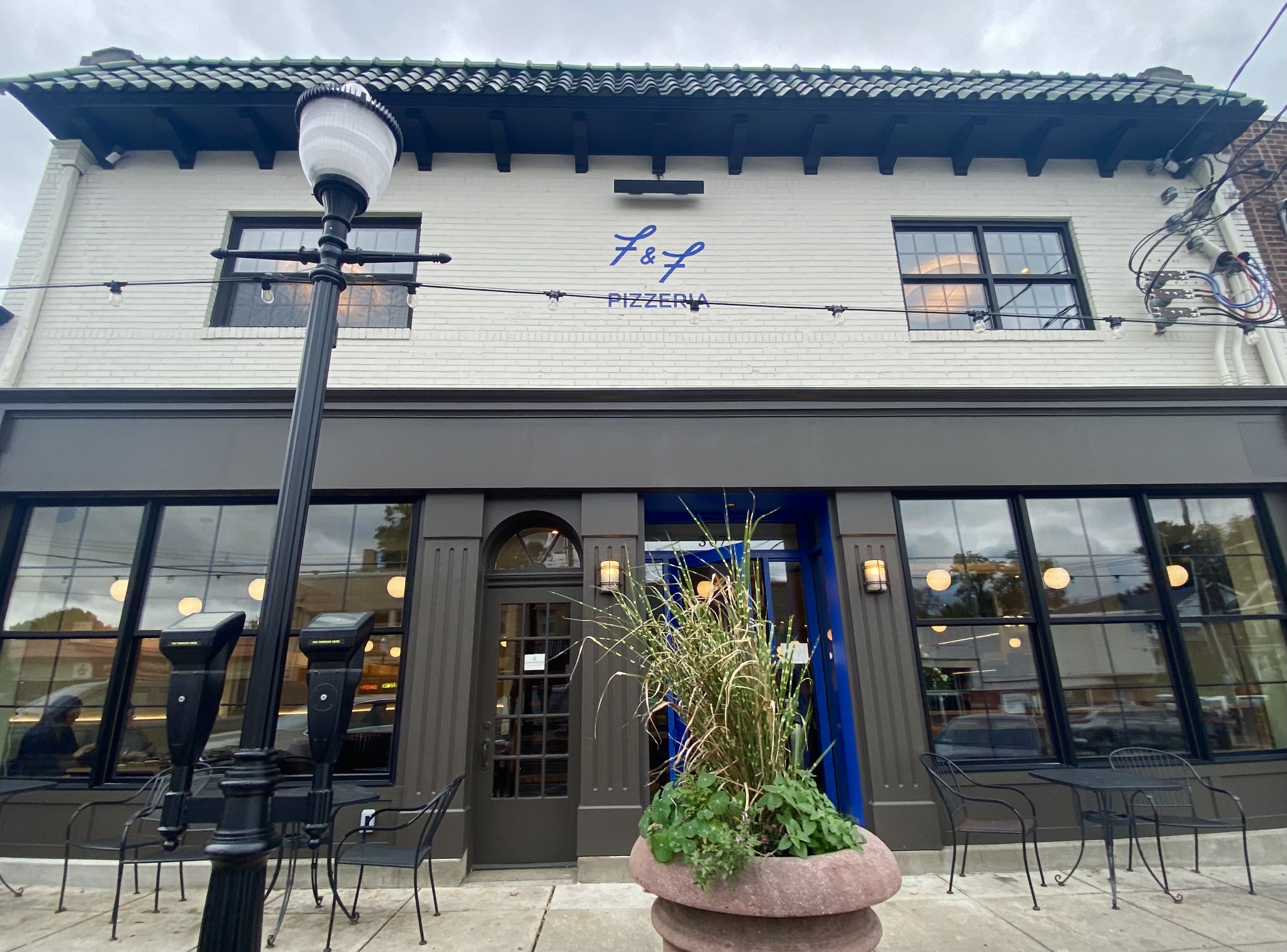 Exterior of F&F Pizzeria with white upper wall, dark lower facade, green-tiled roof, large windows, black outdoor chairs, a streetlamp, and a large planter with tall grass.