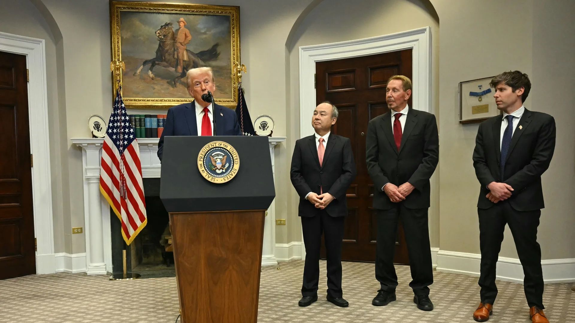 Photo of President Trump at podium. Masayoshi Son, Larry Ellison and Sam Altman stand next to him in black suits.
