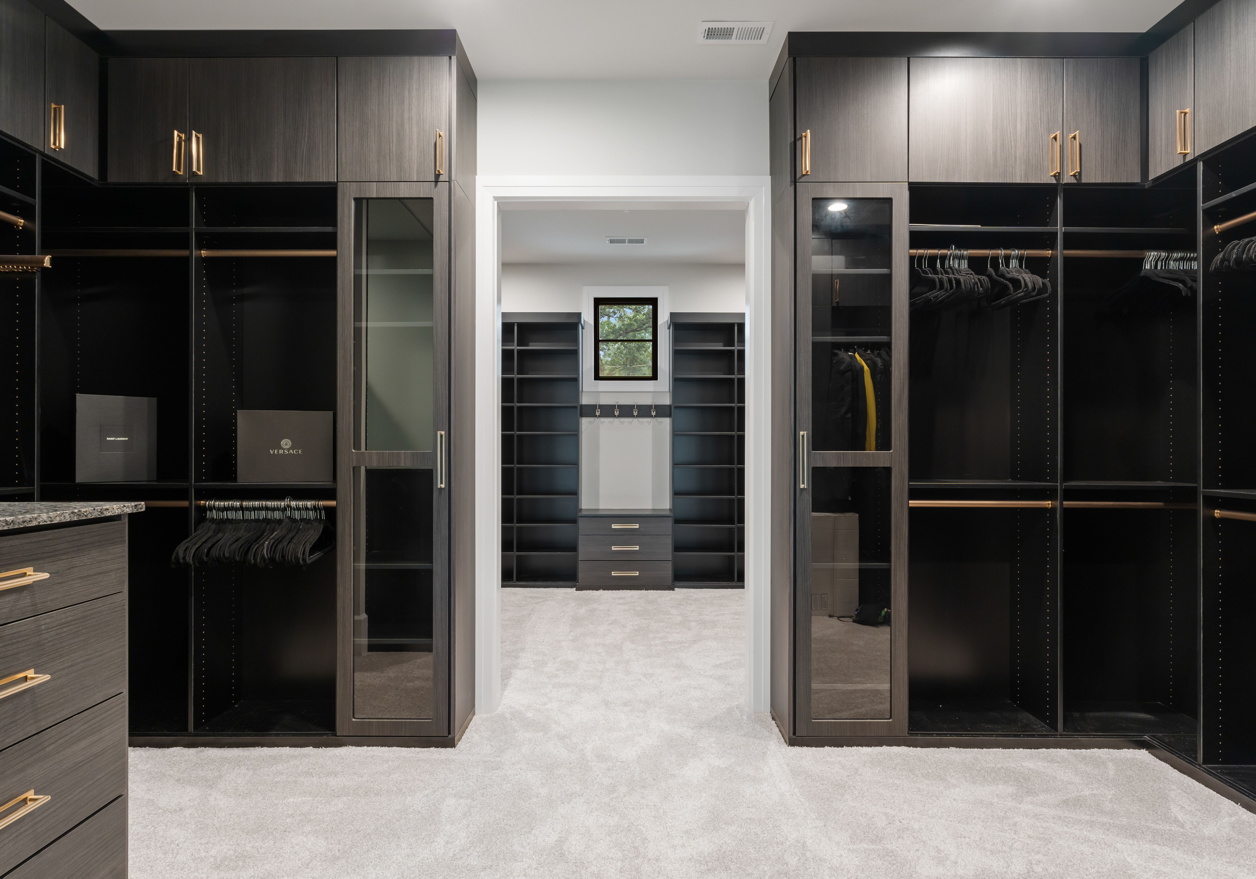 Modern, spacious walk-in closet with dark wood cabinetry, gold handles, black hangers, and light gray carpet. Shelving and drawers visible through central doorway with window above.