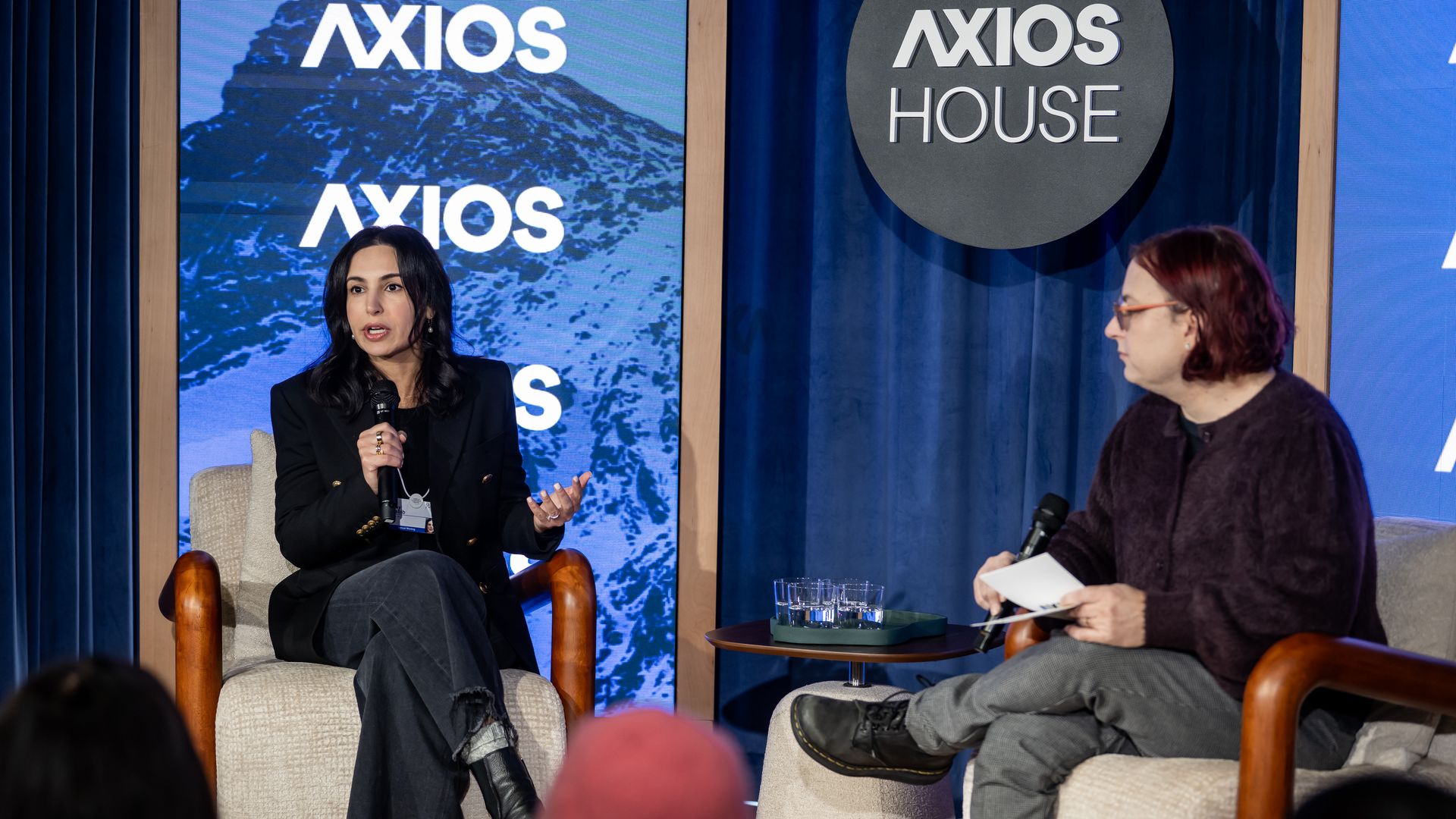Two people speaking on stage at an Axios House event. The person on the left, in a black blazer and jeans, is holding a microphone. The person on the right has red hair and glasses.