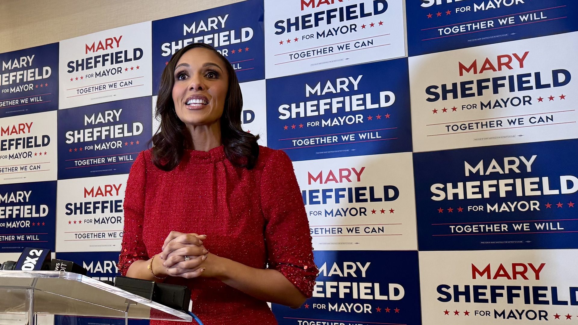 Mary Sheffield speaking at a podium with a backdrop of blue and white campaign signs reading "Mary Sheffield for Mayor" and slogans "Together We Will" and "Together We Can".