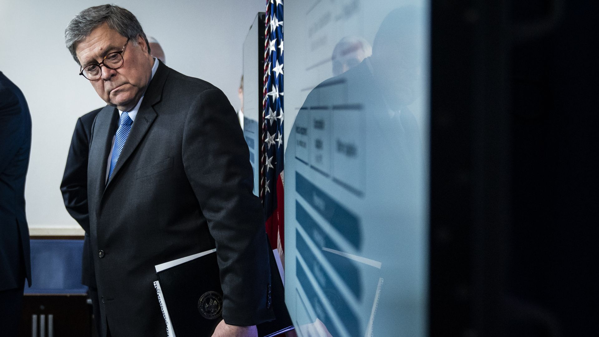 In this image, Barr holds a folder while looking at a TV screen