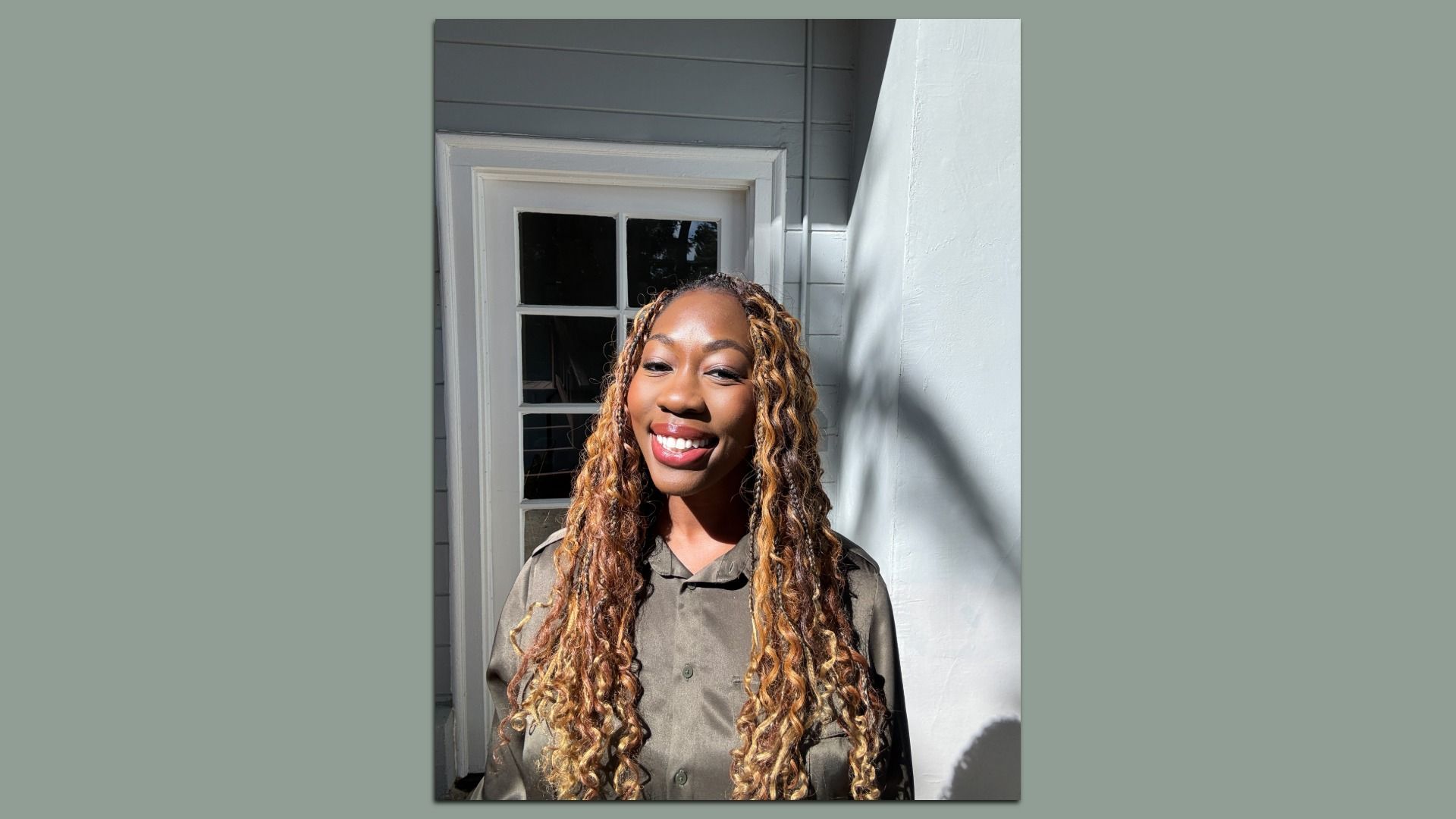 Smiling woman with long curly brown and blonde hair, wearing a green button-up shirt, standing in front of a white door, sunlight casting shadows on the wall.