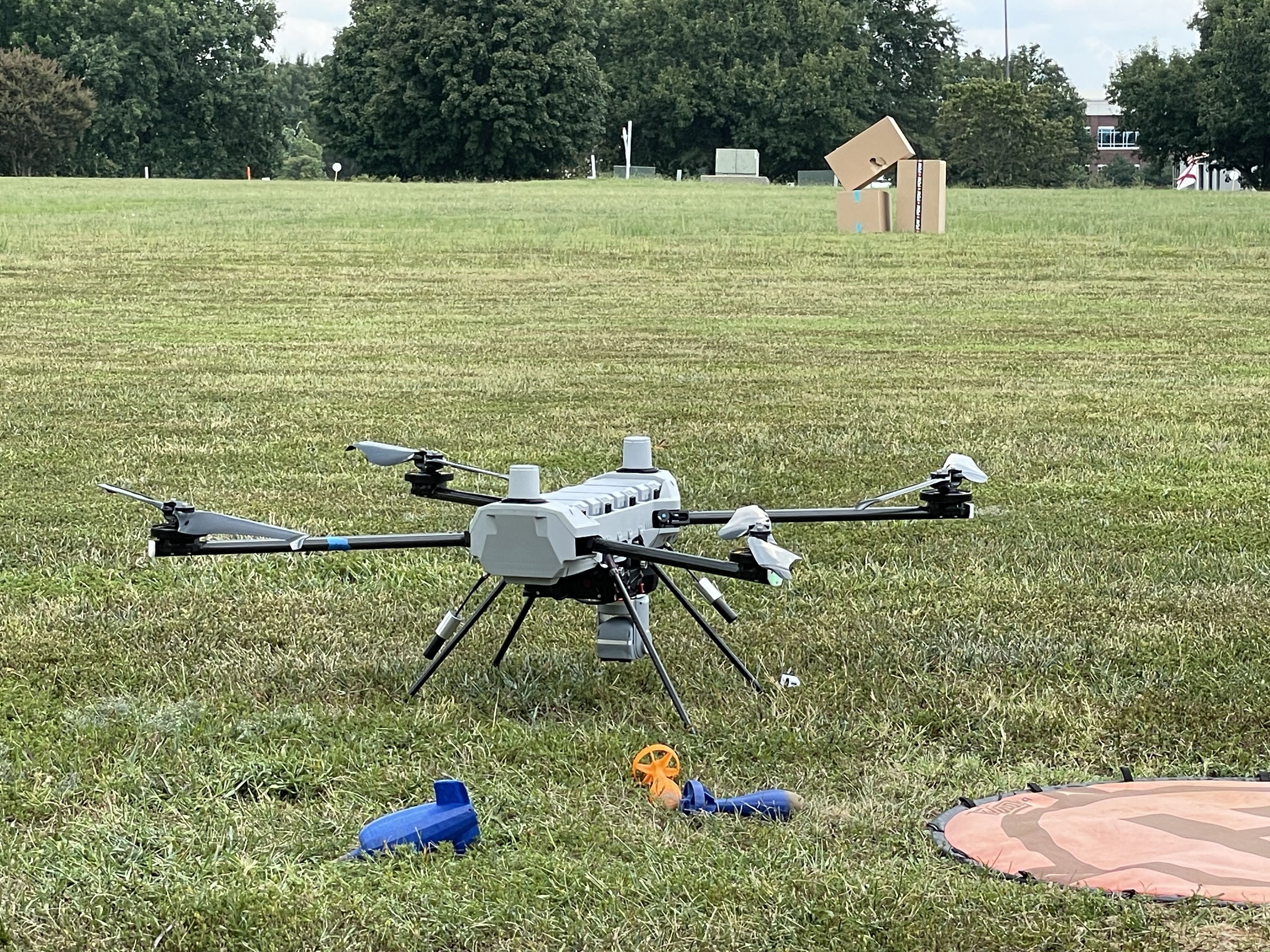 Gray hexacopter drone with six propellers on a grassy field, with blue and orange drone parts nearby and cardboard boxes in the background under a partly cloudy sky.